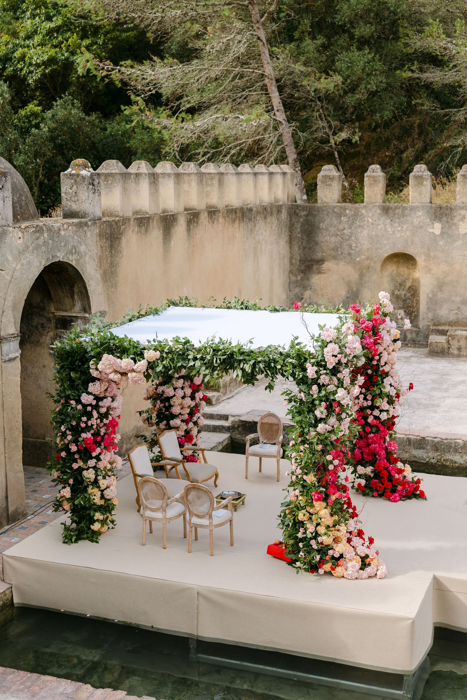 Elegant wedding ceremony setup with white chairs and red floral arrangements against a stone wall backdrop.