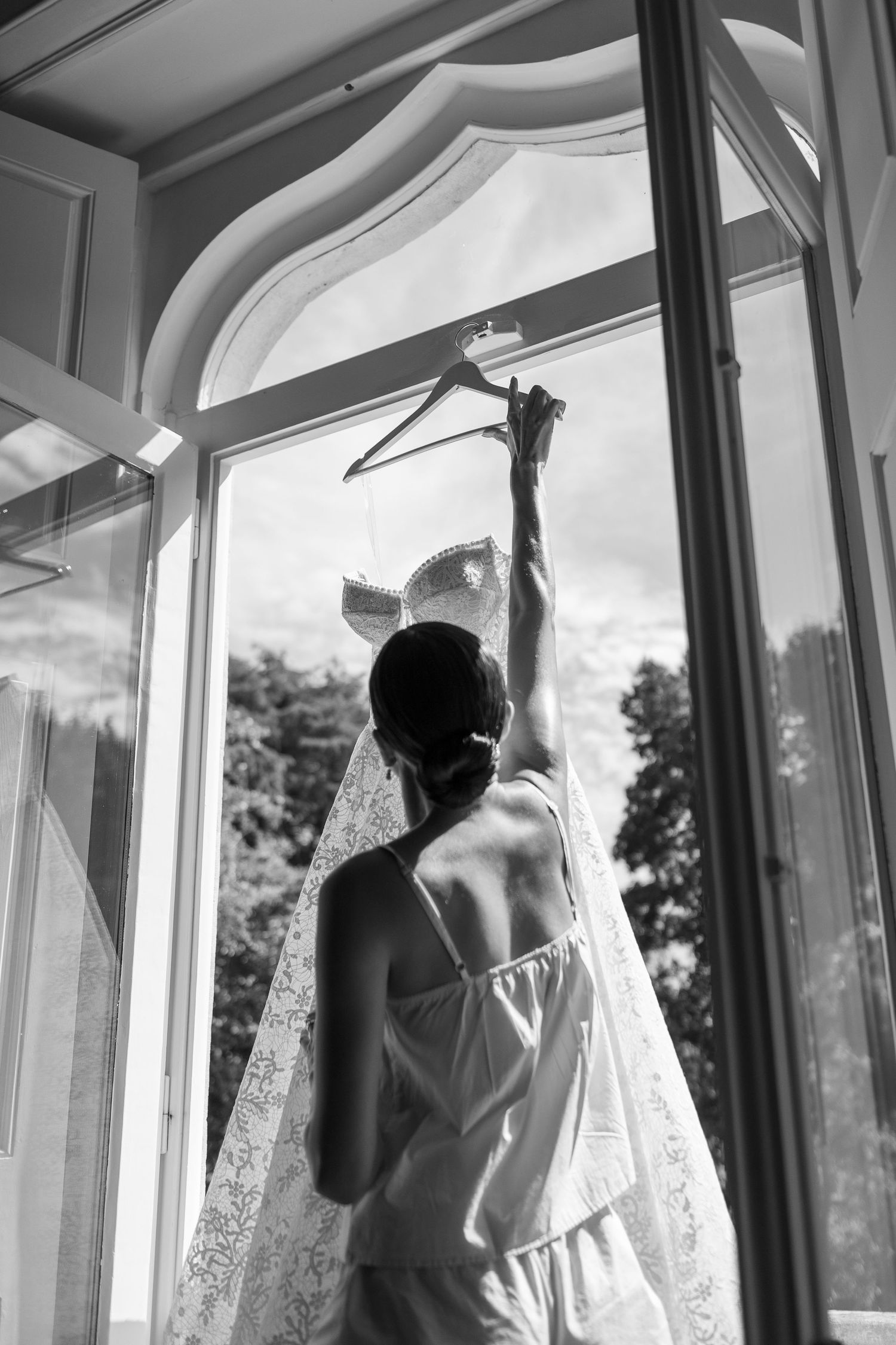 Silhouetted figures admiring a wedding dress hanging by sunlit windows.