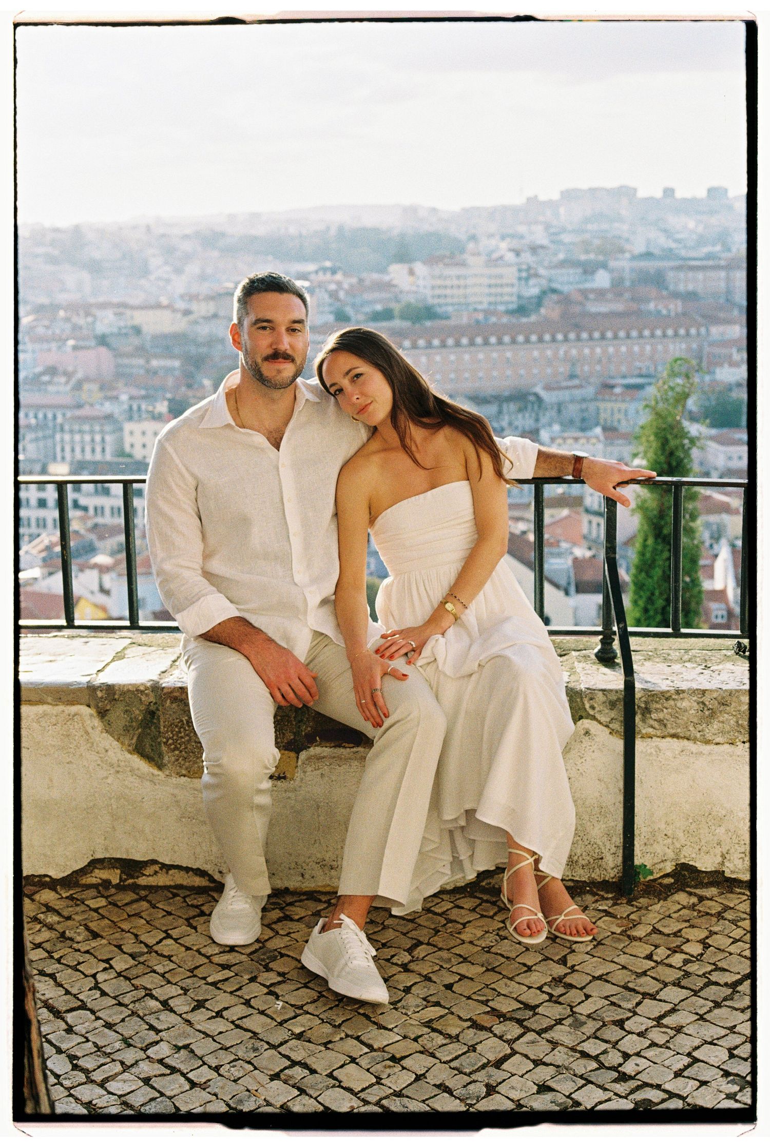 Couple in white outfits sitting on a stone wall with a scenic cityscape backdrop, likely Lisbon, Portugal.
