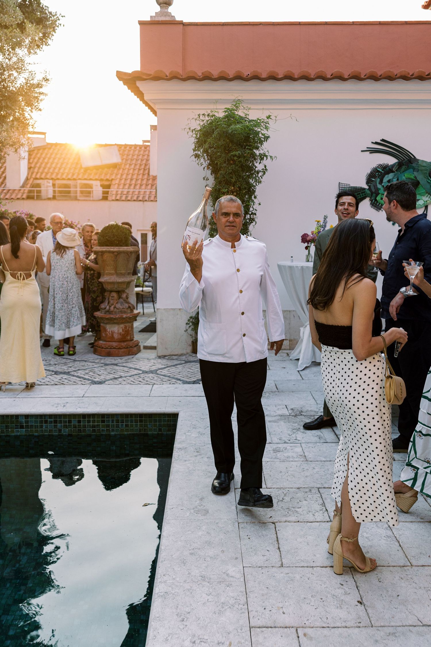 Man in white chef jacket waves at outdoor poolside party during golden hour sunset gathering.