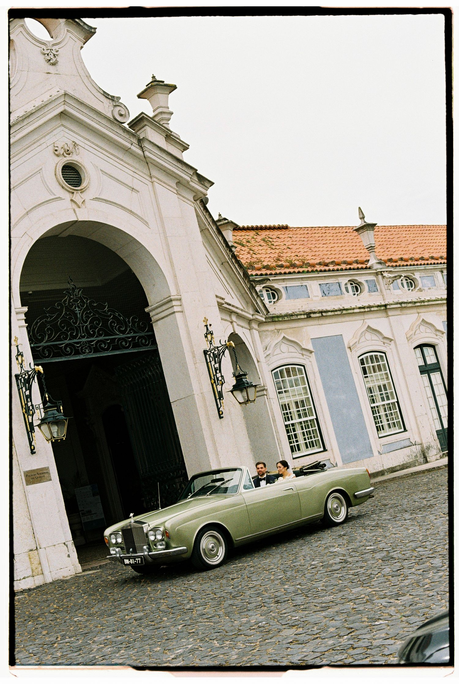 Vintage green Rolls-Royce convertible parked outside an elegant white European palace with ornate arch and terracotta roof.