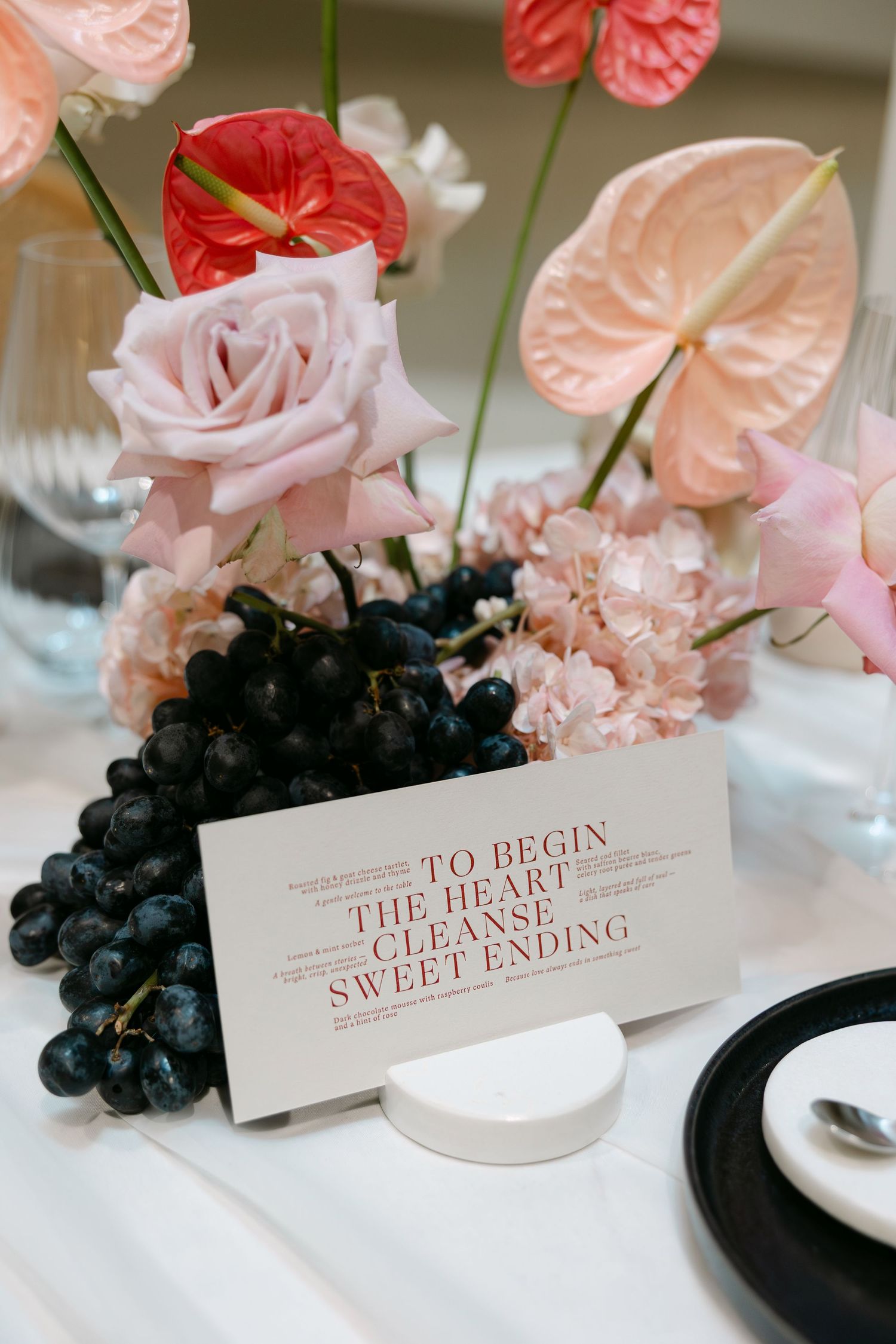 Pink roses, red anthuriums, and dark grapes surround a menu card reading To Begin, The Heart, Cleanse, Sweet Ending.