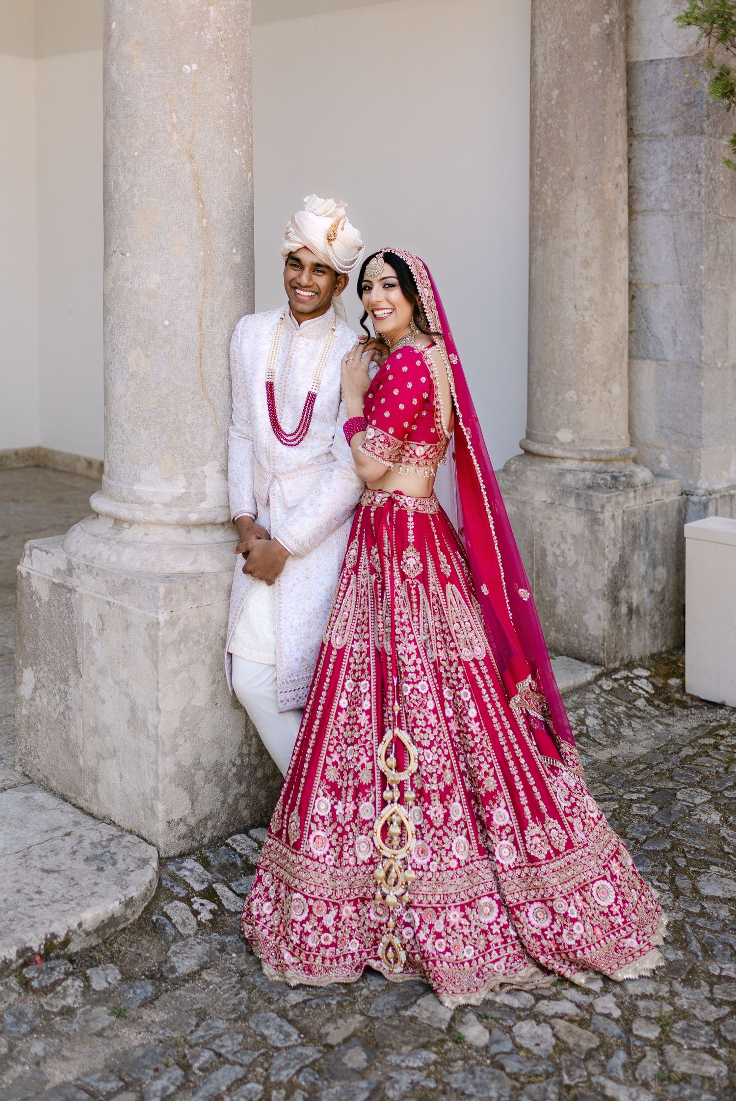 Traditional Indian wedding couple in red and white attire posing against stone pillars.
