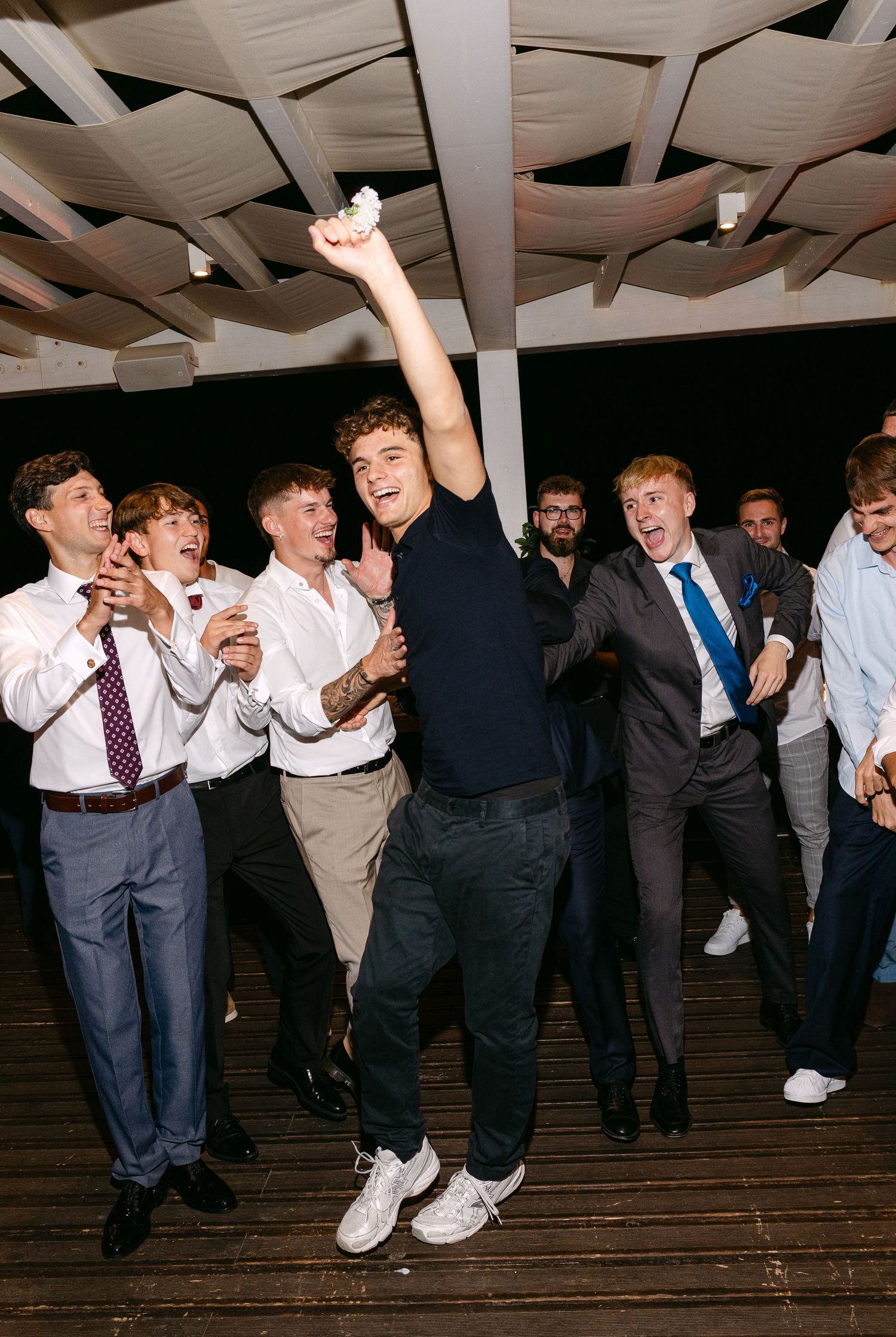 Energetic wedding guests dancing and celebrating at a reception with one person jumping enthusiastically in the center.