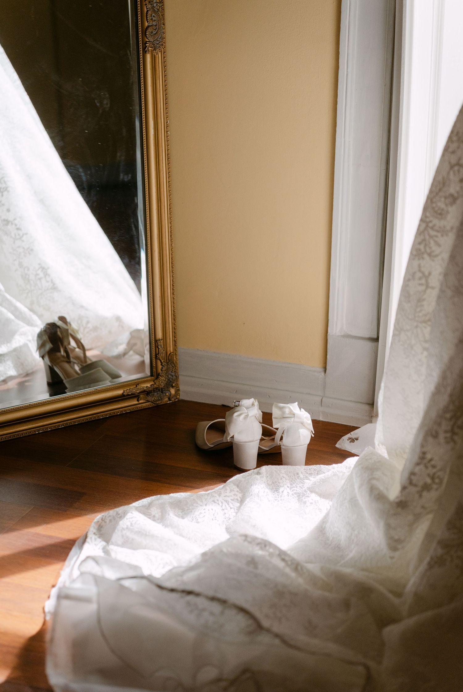 White wedding shoes and dress hem next to an ornate gold mirror on hardwood floor with sunlight streaming in.