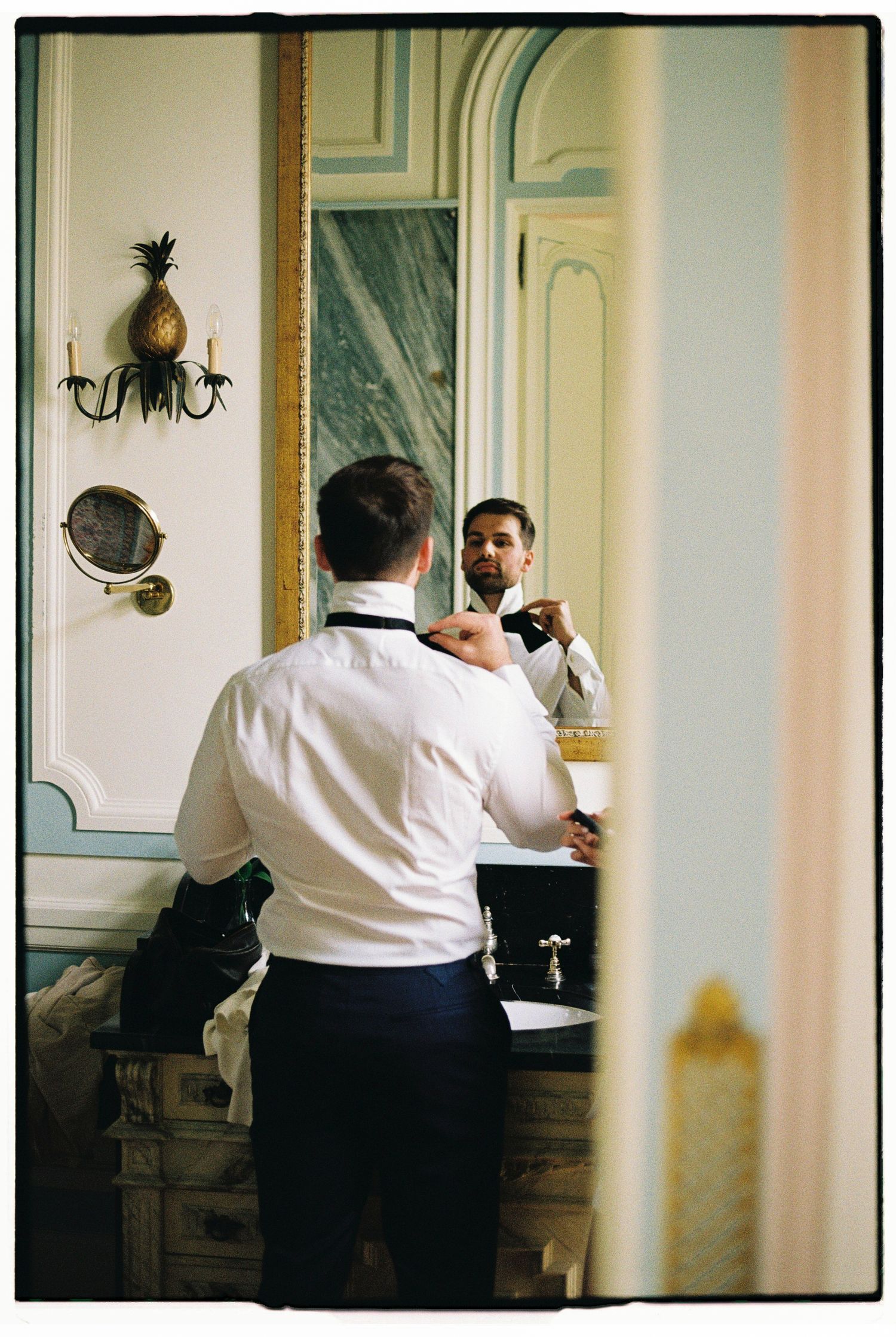 Groom adjusting his bow tie while looking in an ornate mirror in an elegant bathroom before his wedding.