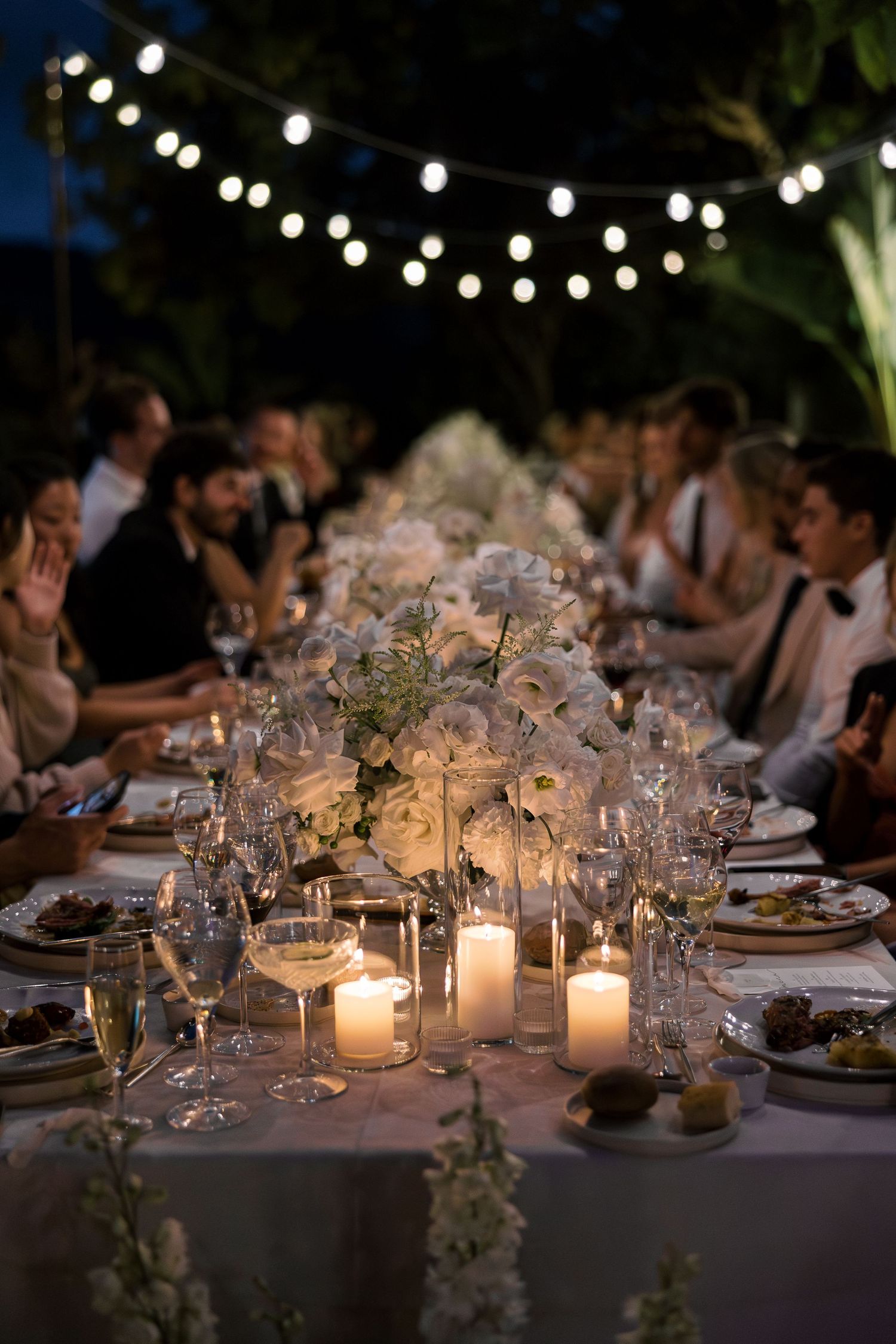 Elegant outdoor wedding reception with candlelit long table, white floral centerpieces, and string lights overhead at night.