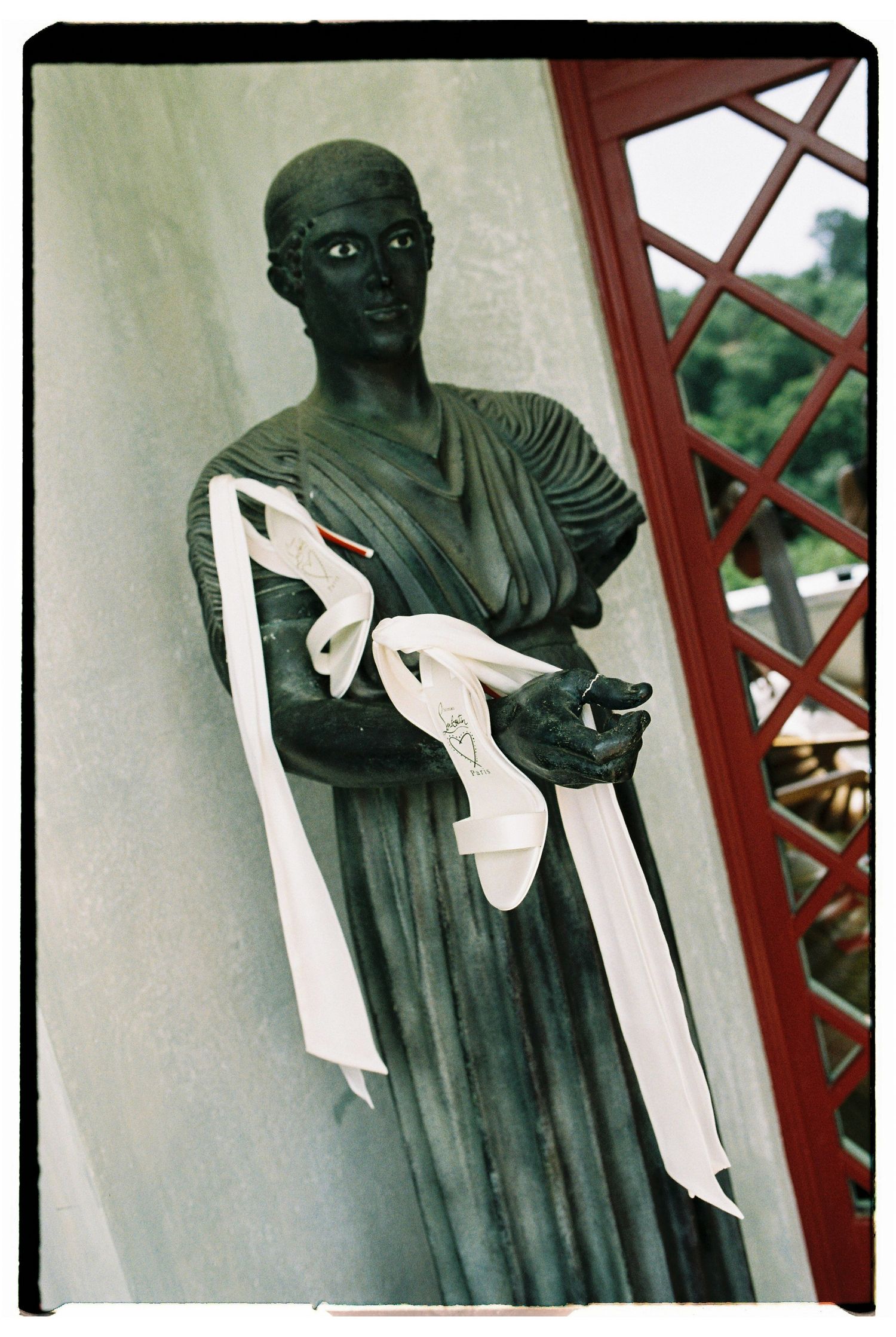 Bronze classical statue adorned with white ribbon sash near red lattice window.