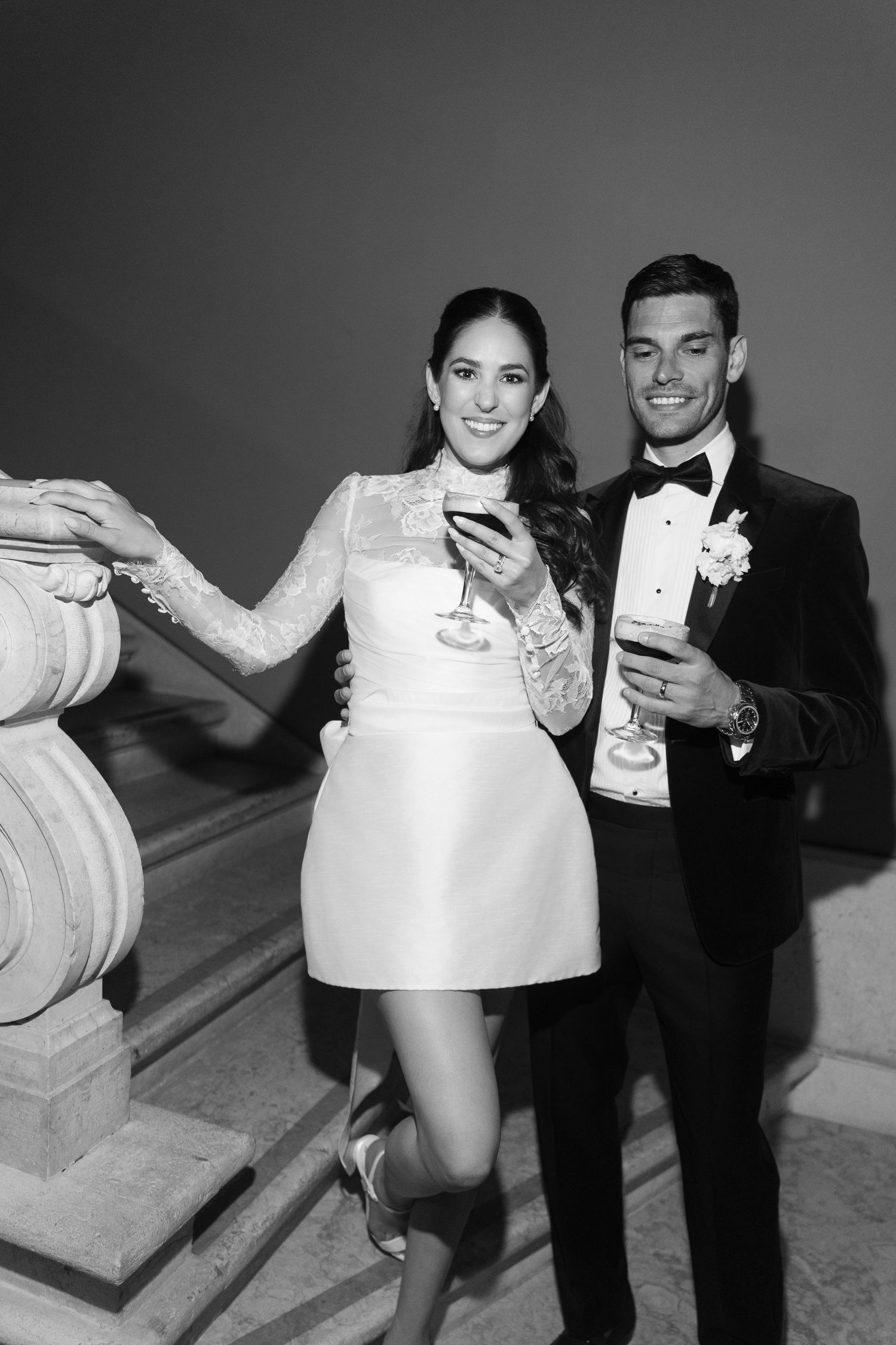 Black and white photo of smiling bride in mini dress and groom in tuxedo holding champagne glasses.