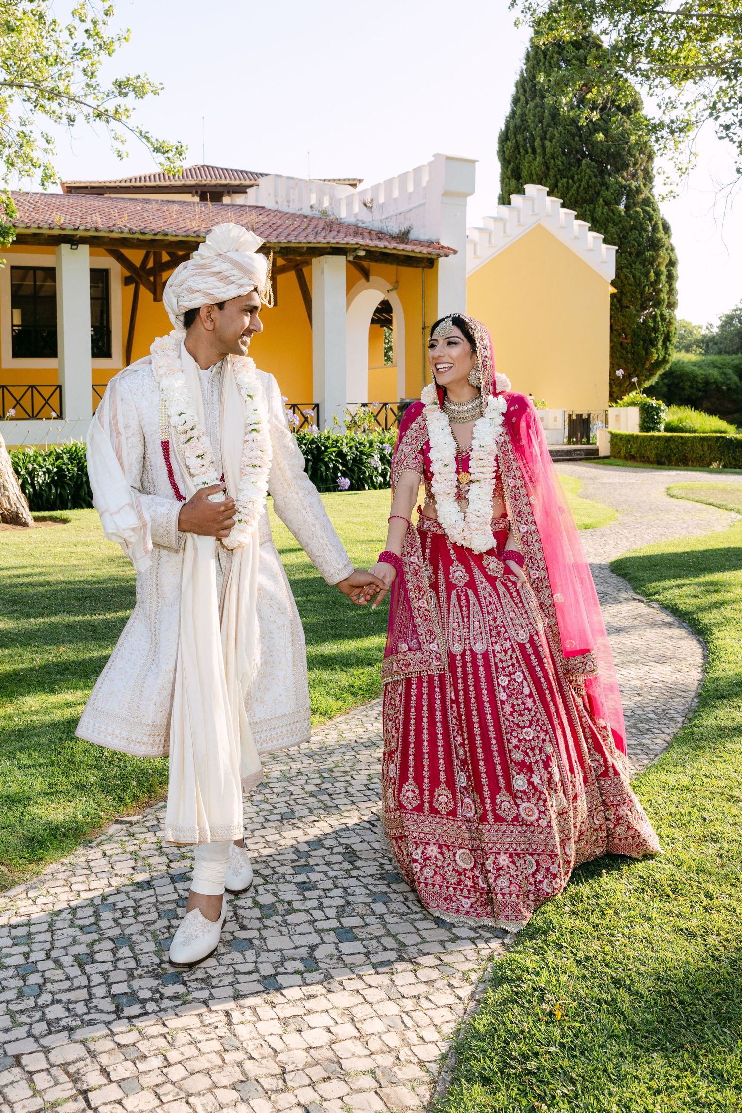 Couple in traditional Indian wedding attire walking together on cobblestone path near yellow buildings.