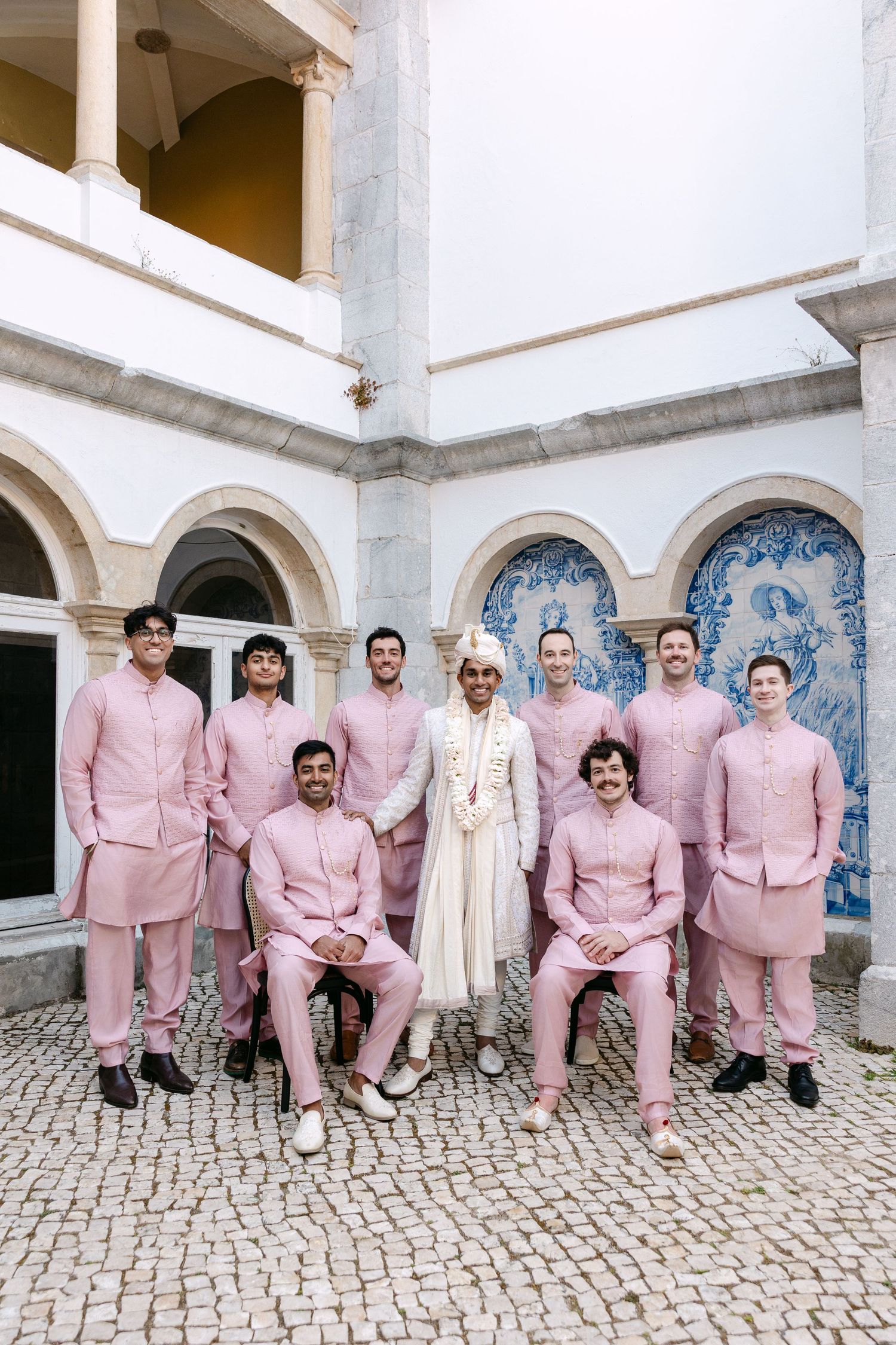 Wedding party members in matching pink outfits pose against white building with blue tile accents.