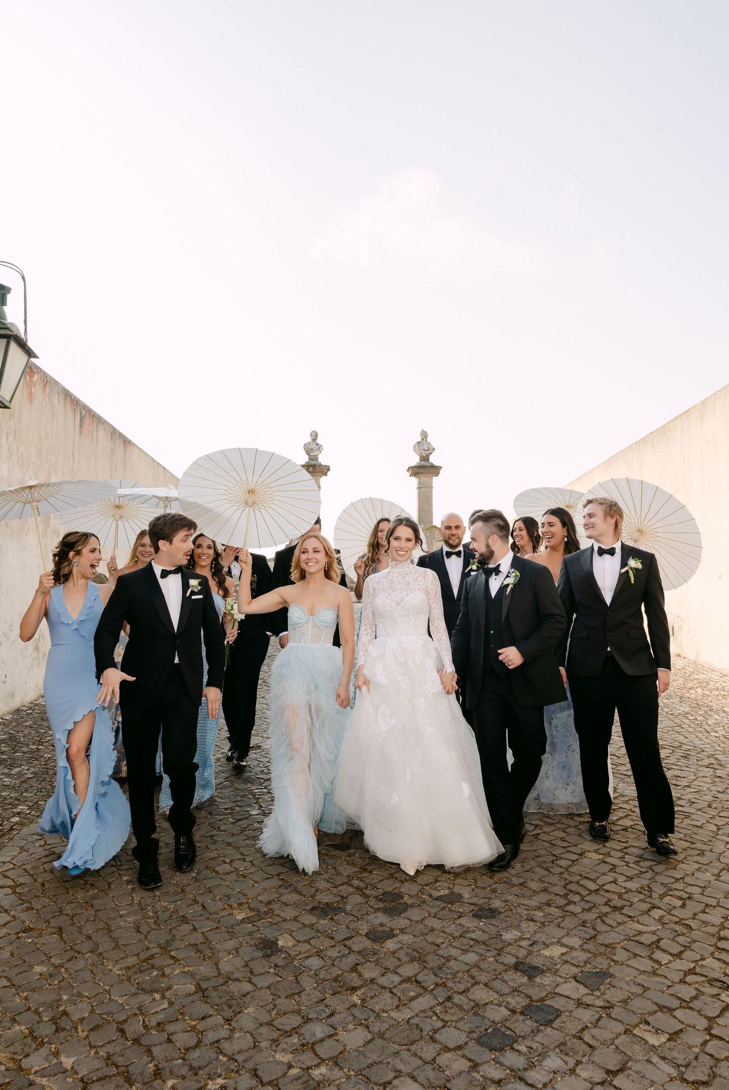 Wedding party walking together with parasols along cobblestone path at sunset.