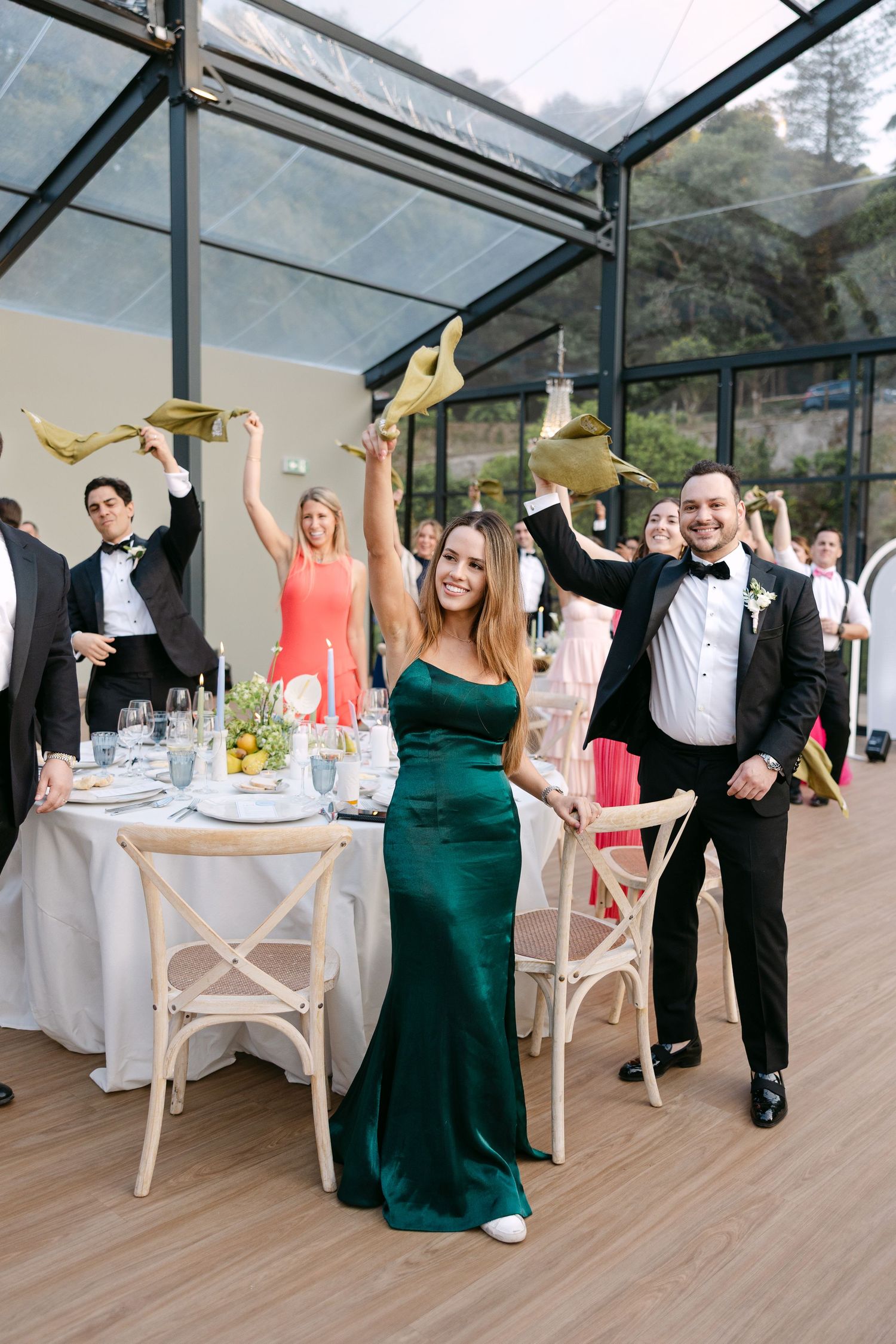 Wedding guests celebrating with raised arms in a glass-ceiling reception venue.