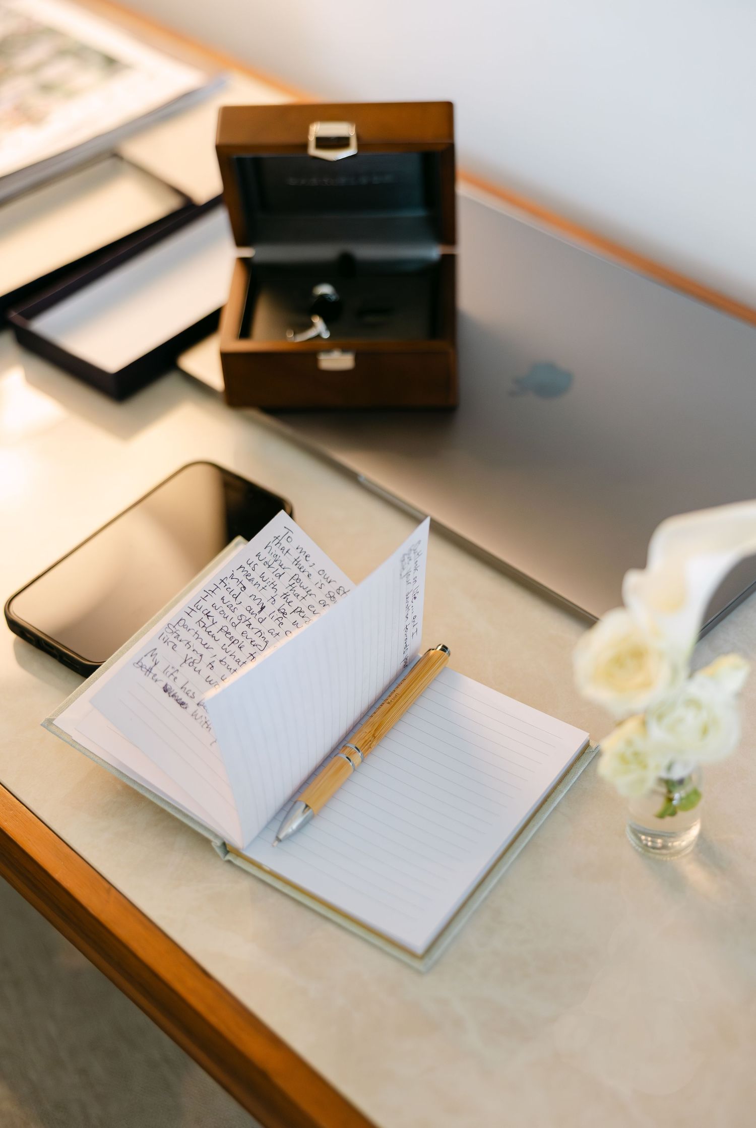 Wedding details with open notebook, pen and wooden jewelry box on desk with white flower.