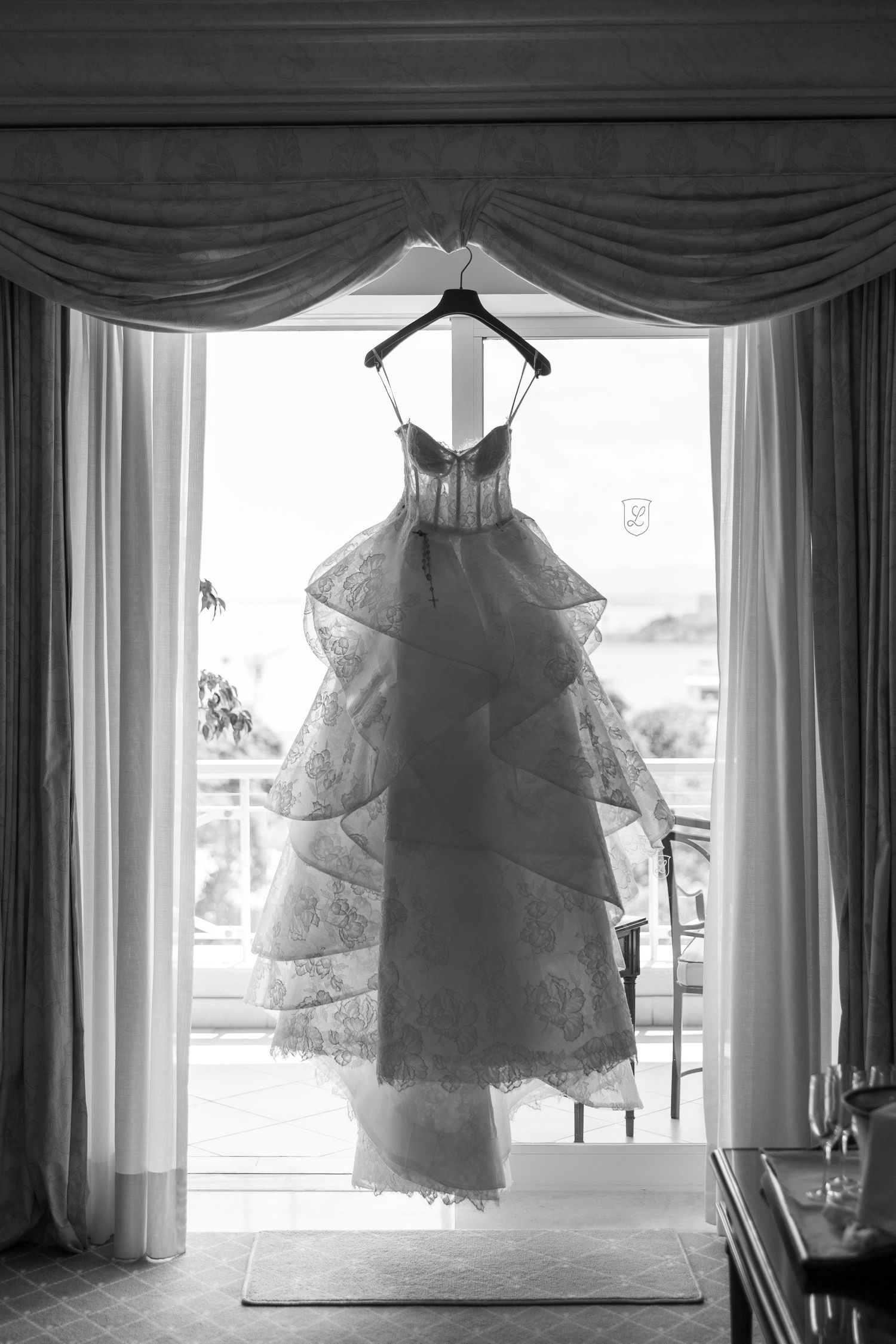 Wedding dress hangs gracefully by window with dramatic lighting and flowing veil.