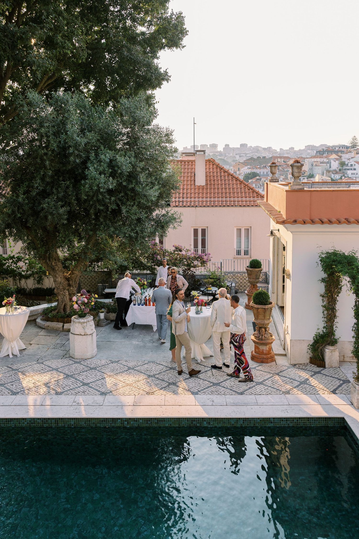 Elegant outdoor garden party near a pool with guests mingling at white-clothed tables at golden hour.
