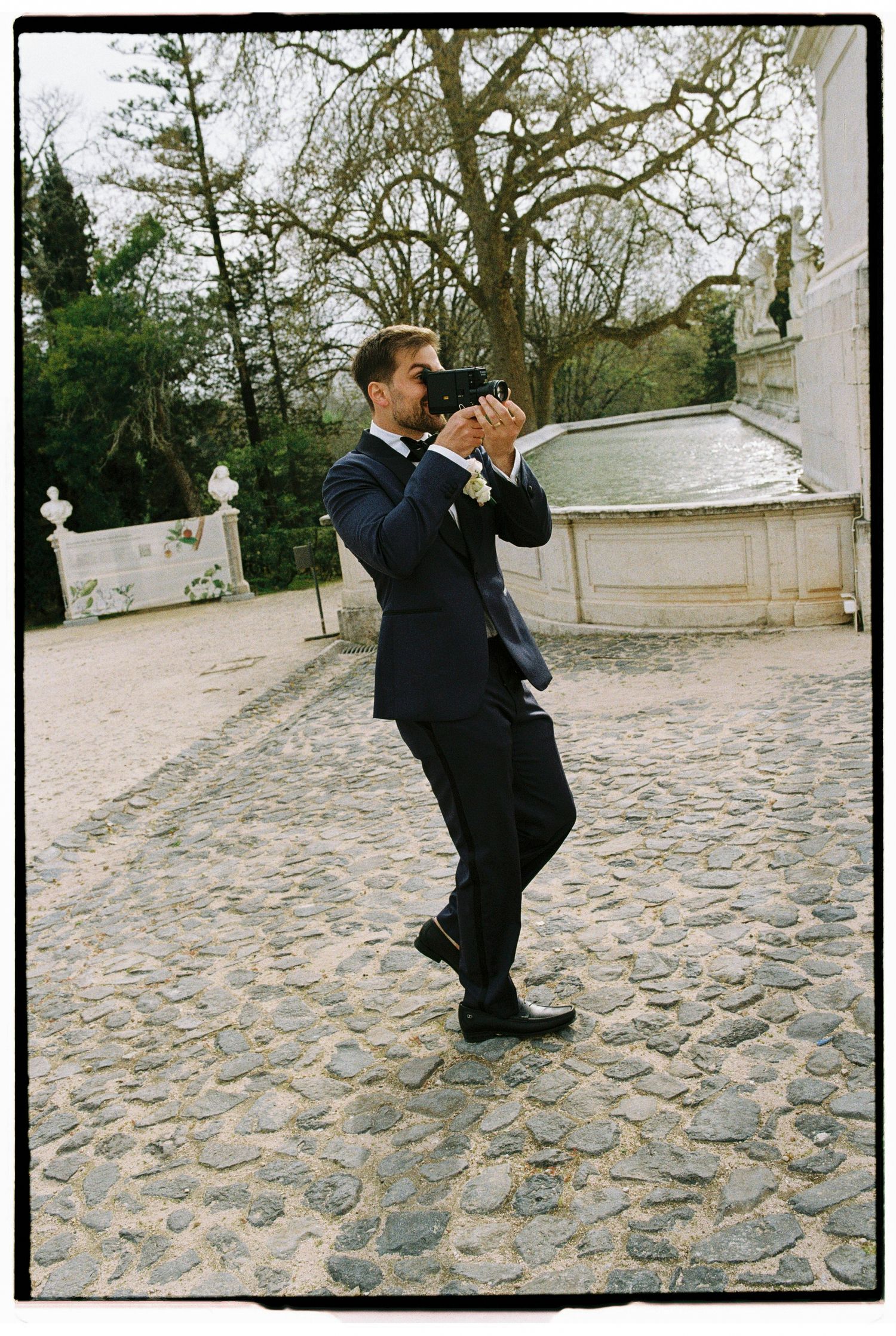 Groom in navy suit photographs with camera on cobblestone path near ornate garden structure.