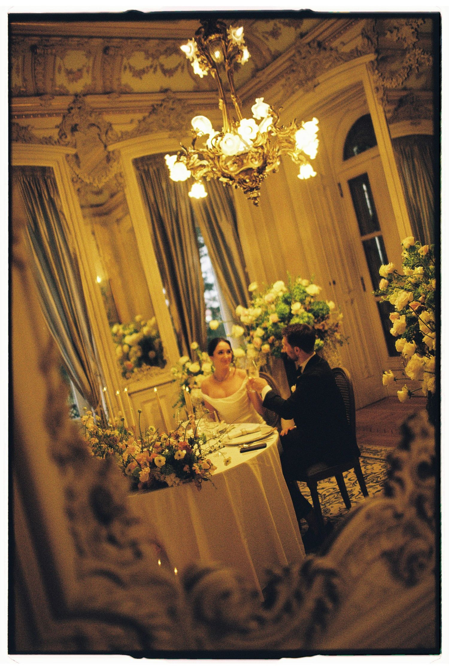 Elegant couple dining in ornate ballroom with chandelier and floral arrangements at a romantic wedding reception.