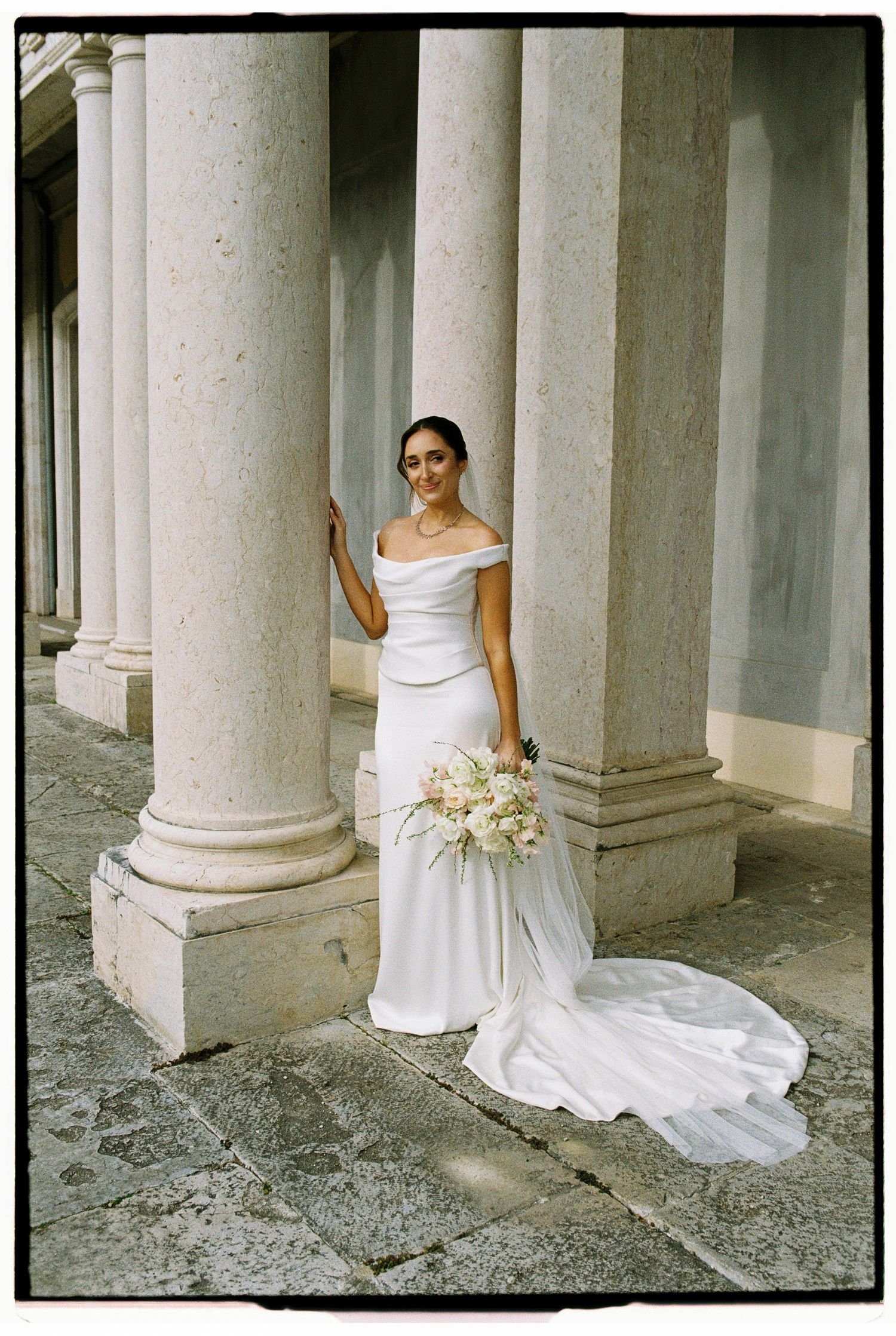 Bride in off-shoulder white gown with train holds bouquet among stone columns.