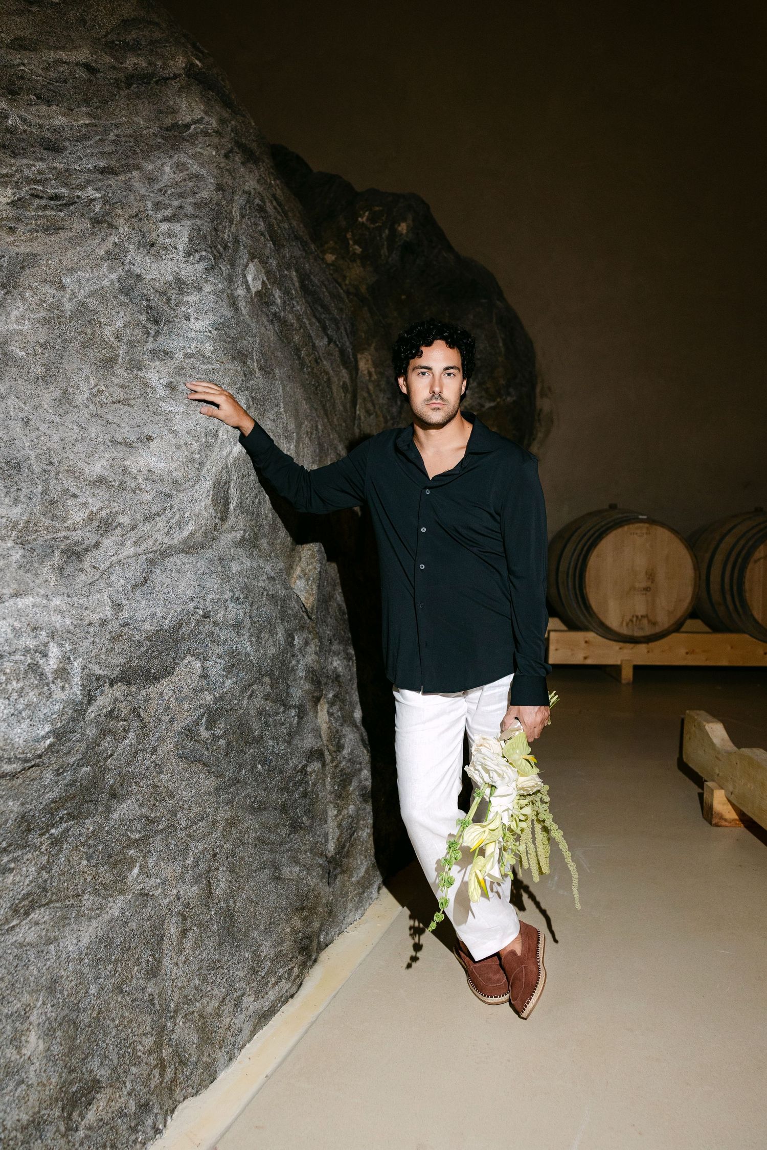 Curly-haired man in black shirt leans against a large rock in a rustic wine cellar with oak barrels.