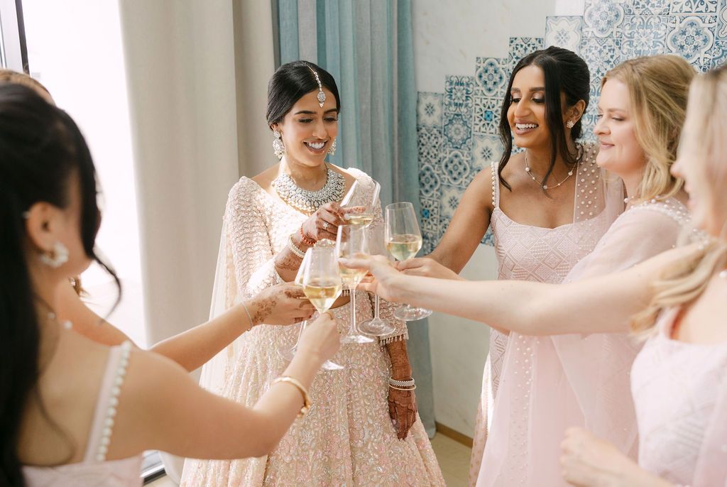 Wedding party members in white dresses toast with champagne glasses during pre-wedding celebrations.