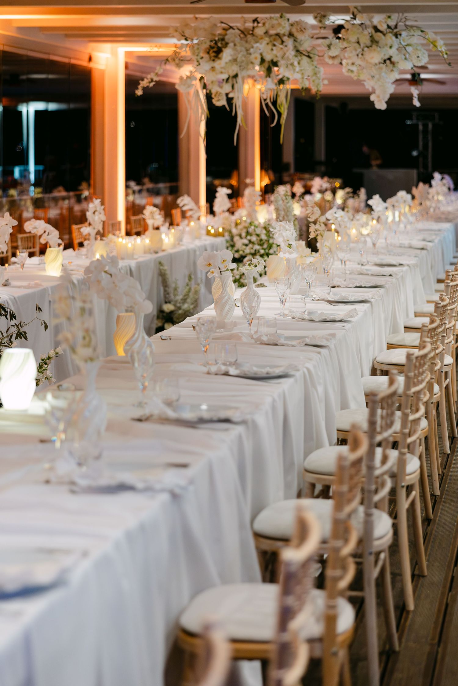 Long banquet tables set with white linens and gold chairs under suspended floral decorations.