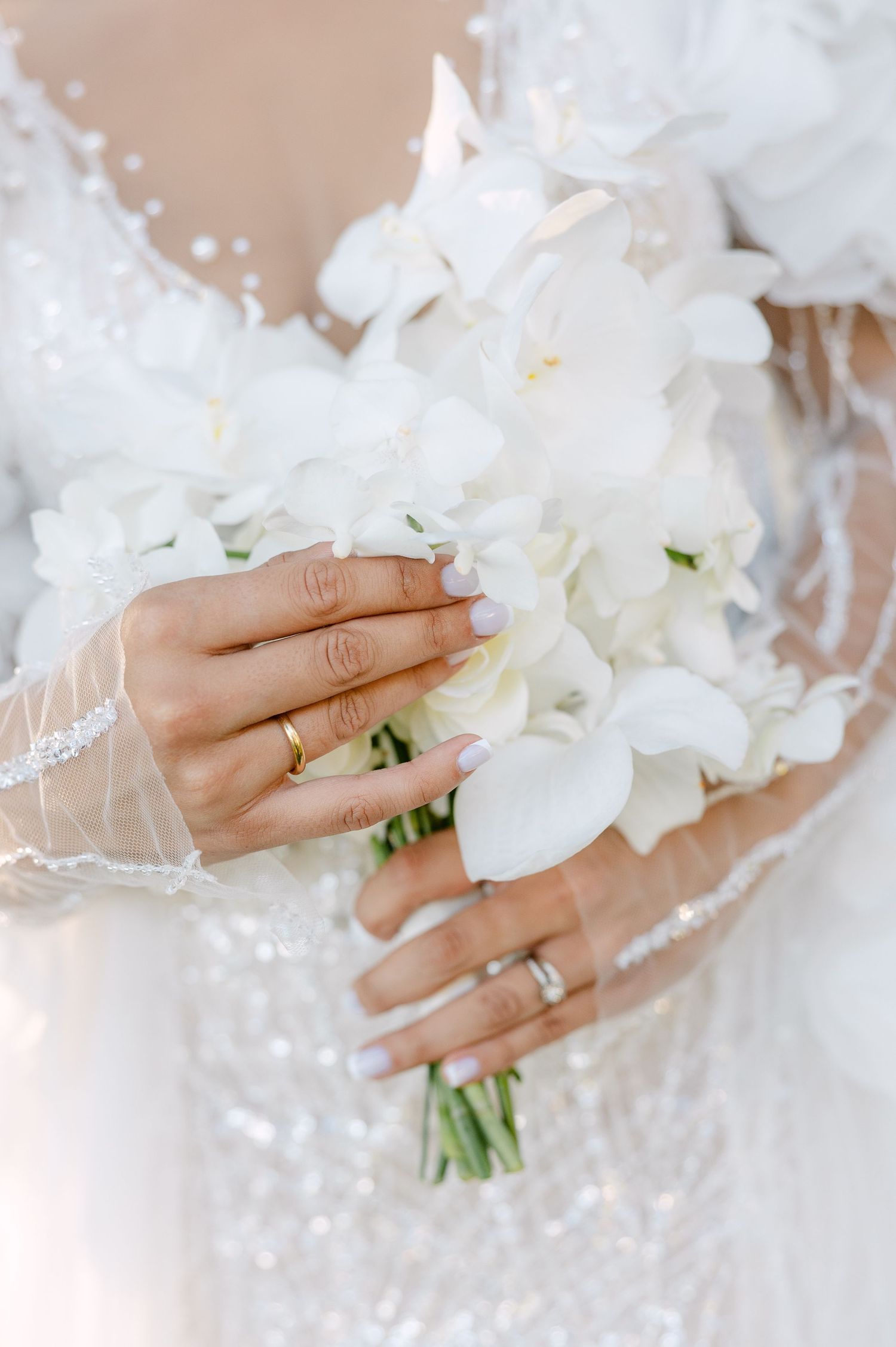 Elegant white wedding bouquet held by hands wearing delicate jewelry and rings.