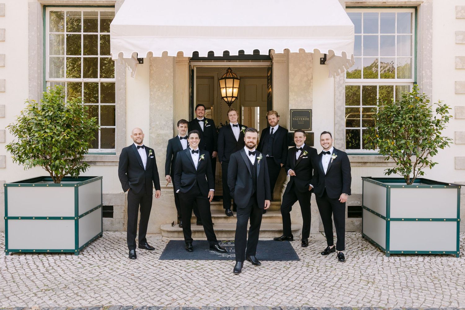 Wedding party poses outside elegant white building with classical architecture.