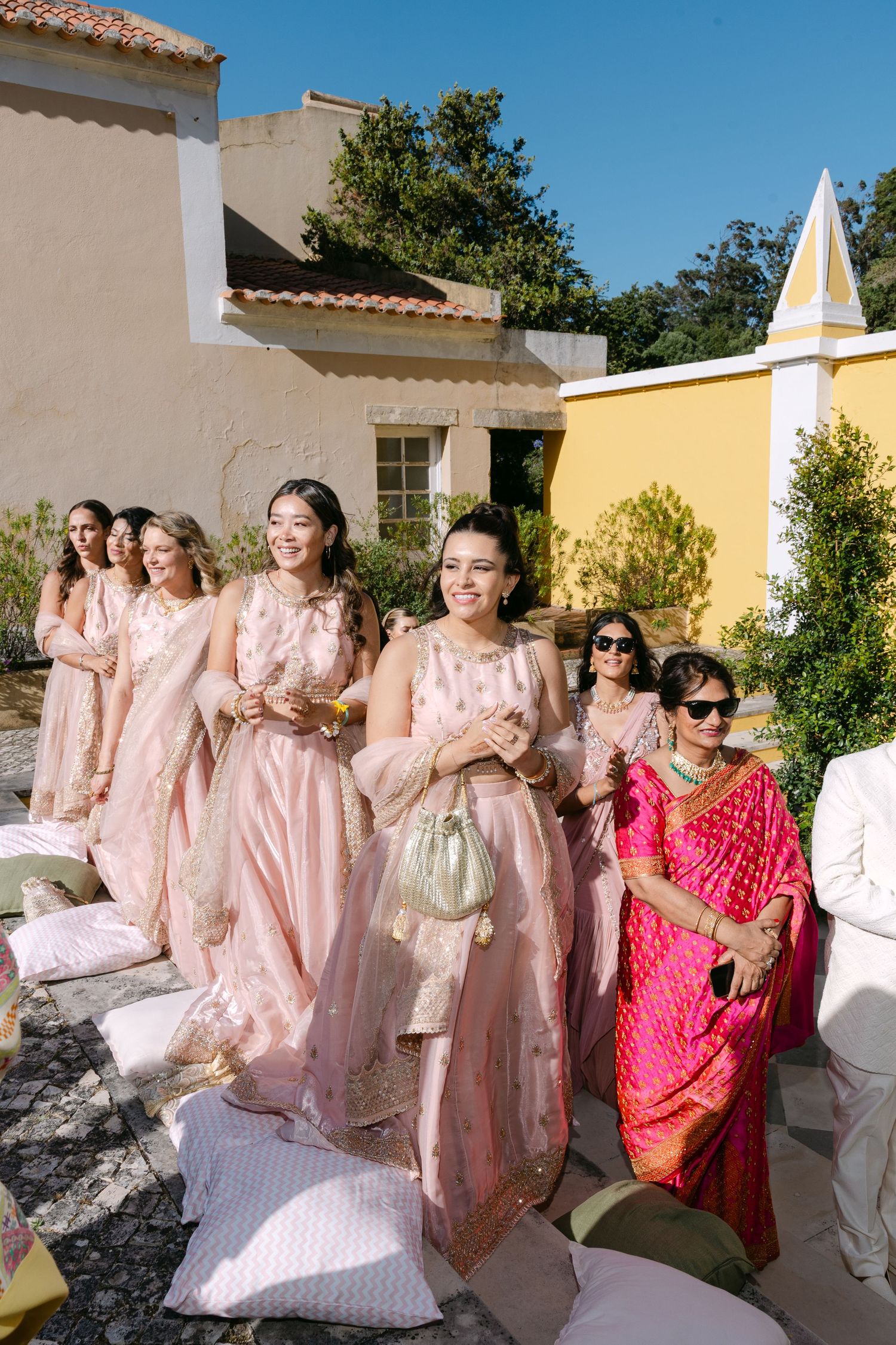 Wedding party in pink dresses and formal attire celebrating outside Mediterranean-style buildings.