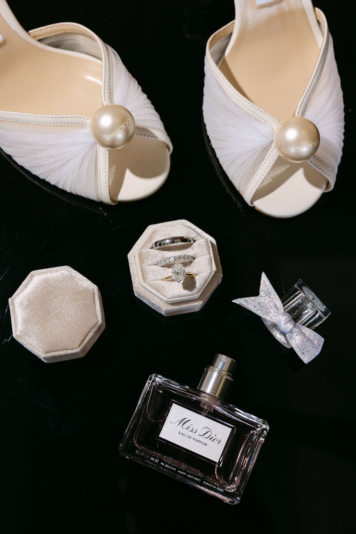Wedding accessories including white bridal shoes and perfume bottle arranged on black background.