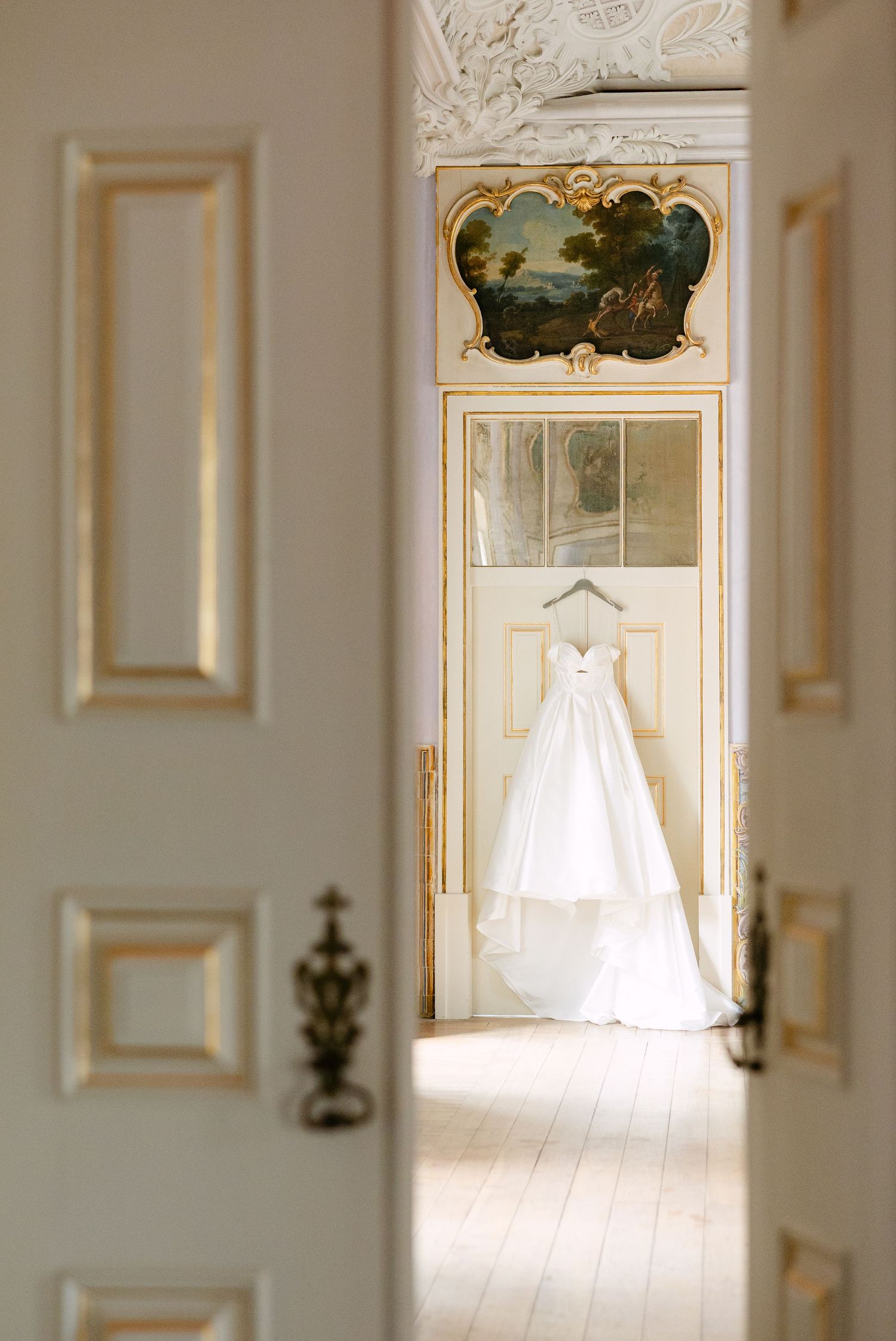 White wedding dress hanging in doorway of elegant room with ornate moldings and natural light.
