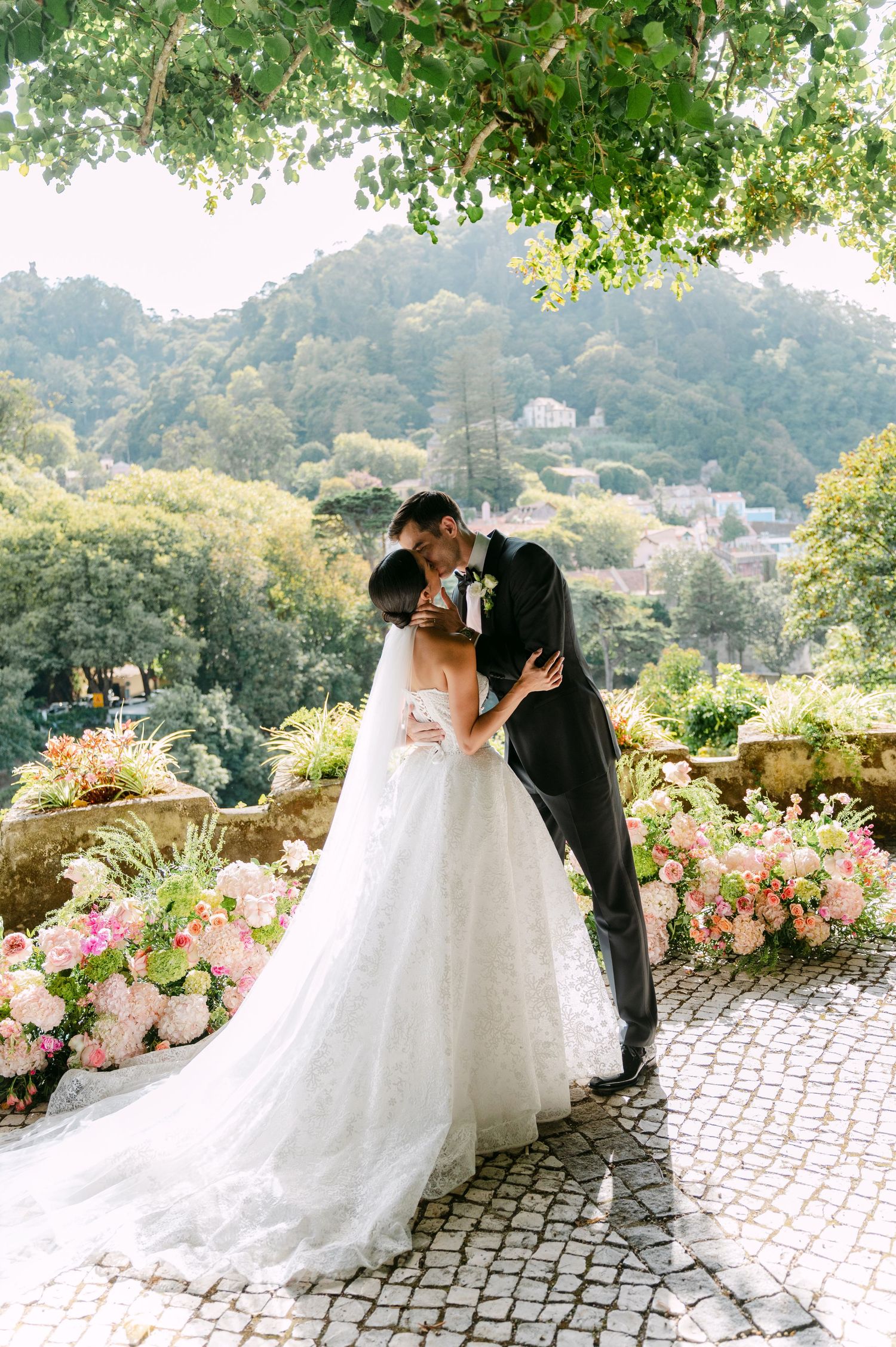 Romantic wedding couple portraits on stone pathway with pink floral arrangements and mountain backdrop.