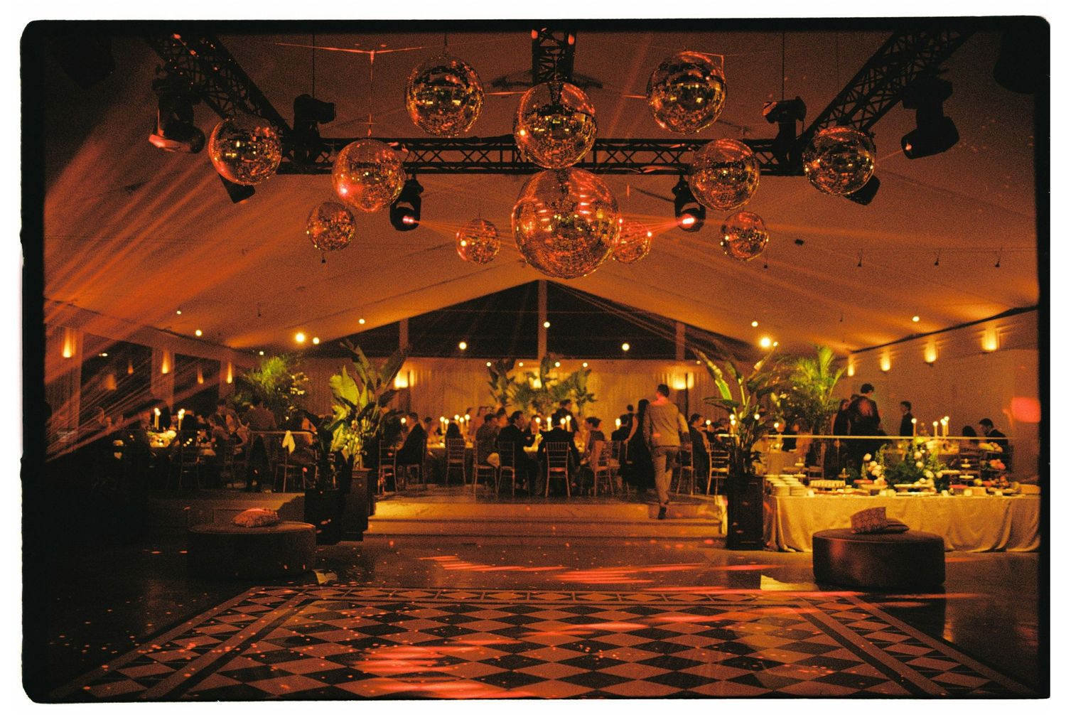 Elegant event tent with disco balls, checkered dance floor, warm amber lighting, and guests mingling at a glamorous evening gala.