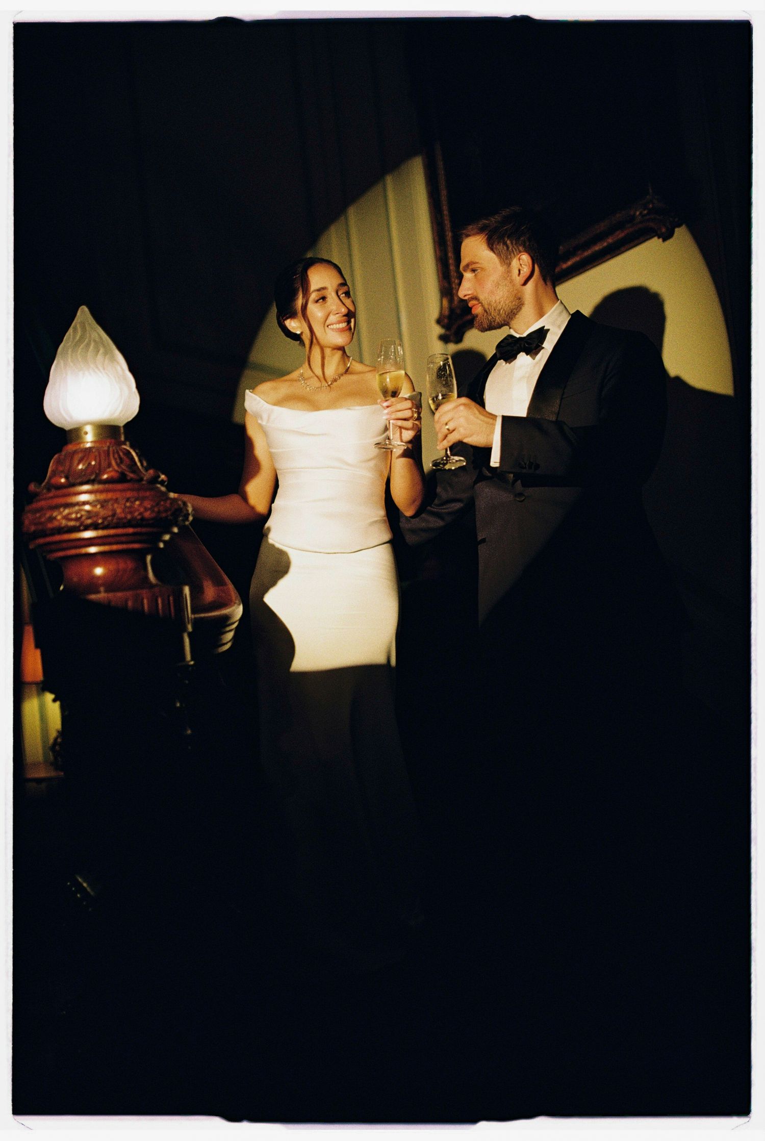Elegant couple in formal attire toasting champagne by a vintage oil lamp in a dimly lit romantic setting.