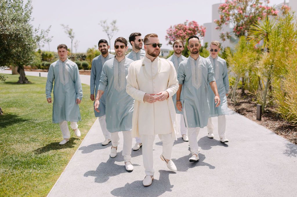 Wedding party members in light blue suits and white attire pose together on a sunny outdoor path.