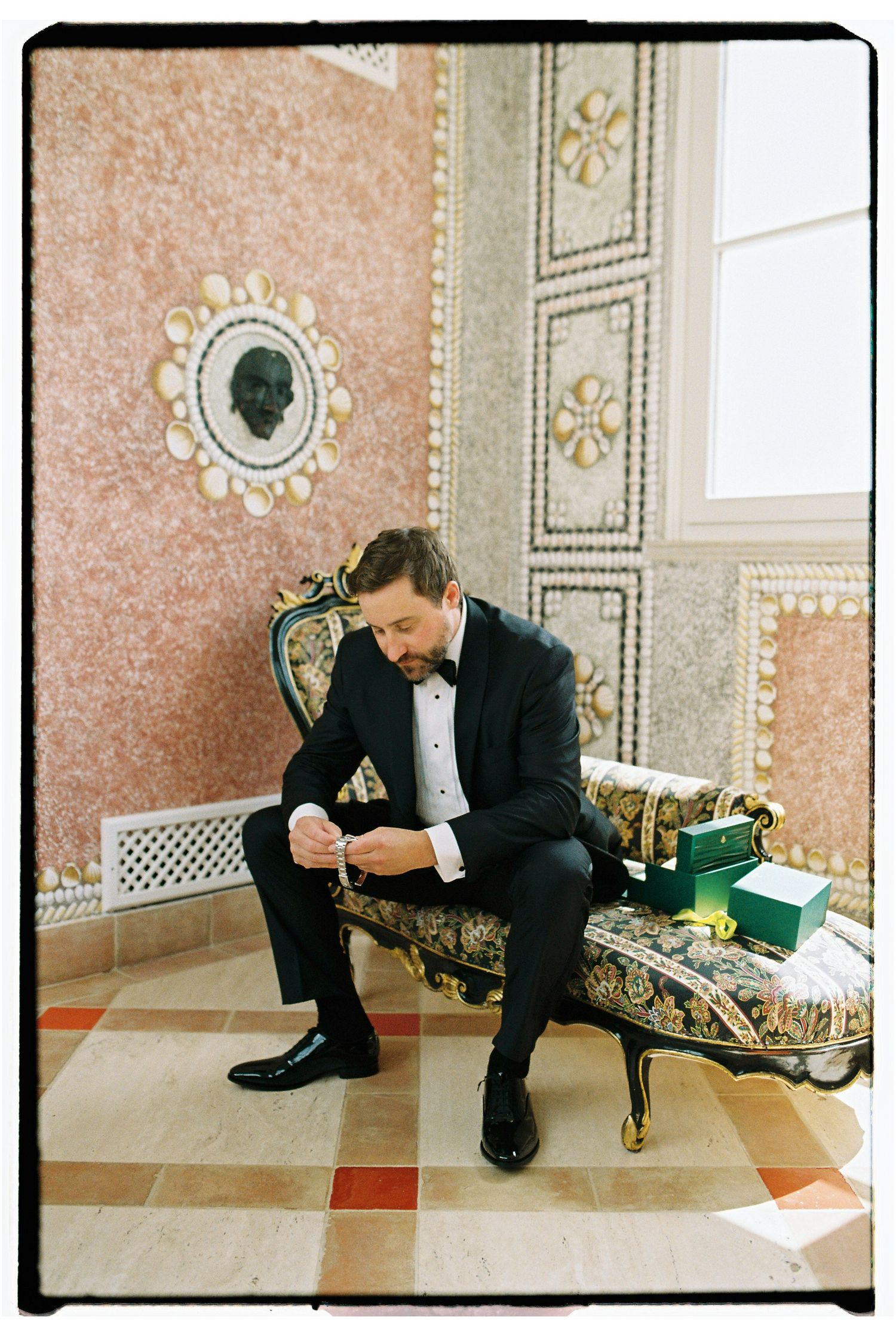 Groom in black tuxedo sits on ornate gold chaise lounge adjusting cufflinks in elegant marble-walled room.