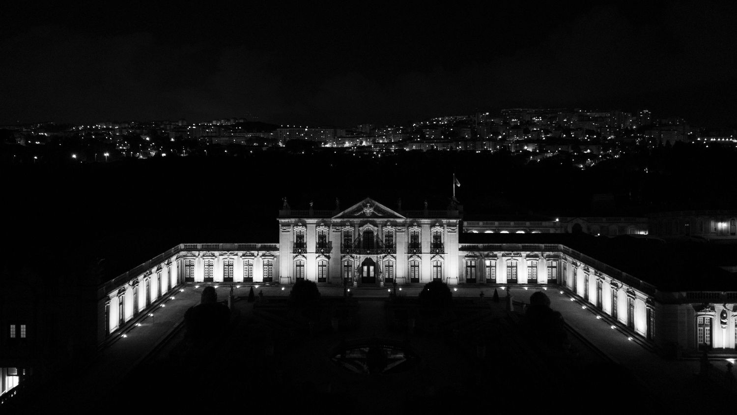 Aerial black and white night view of illuminated grand palace building with city skyline backdrop.