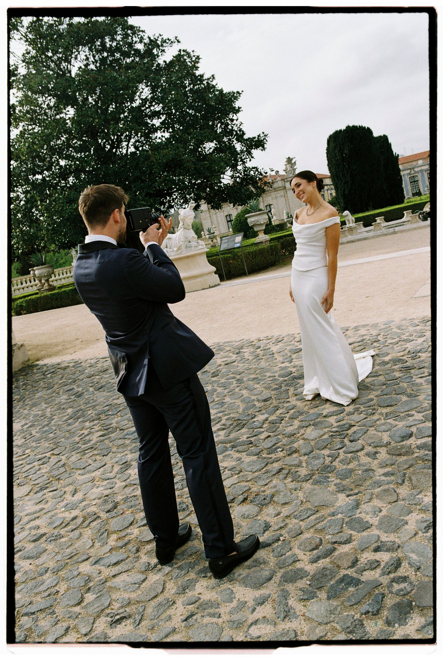 Groom photographs bride in white off-shoulder gown on cobblestone plaza with formal gardens behind.
