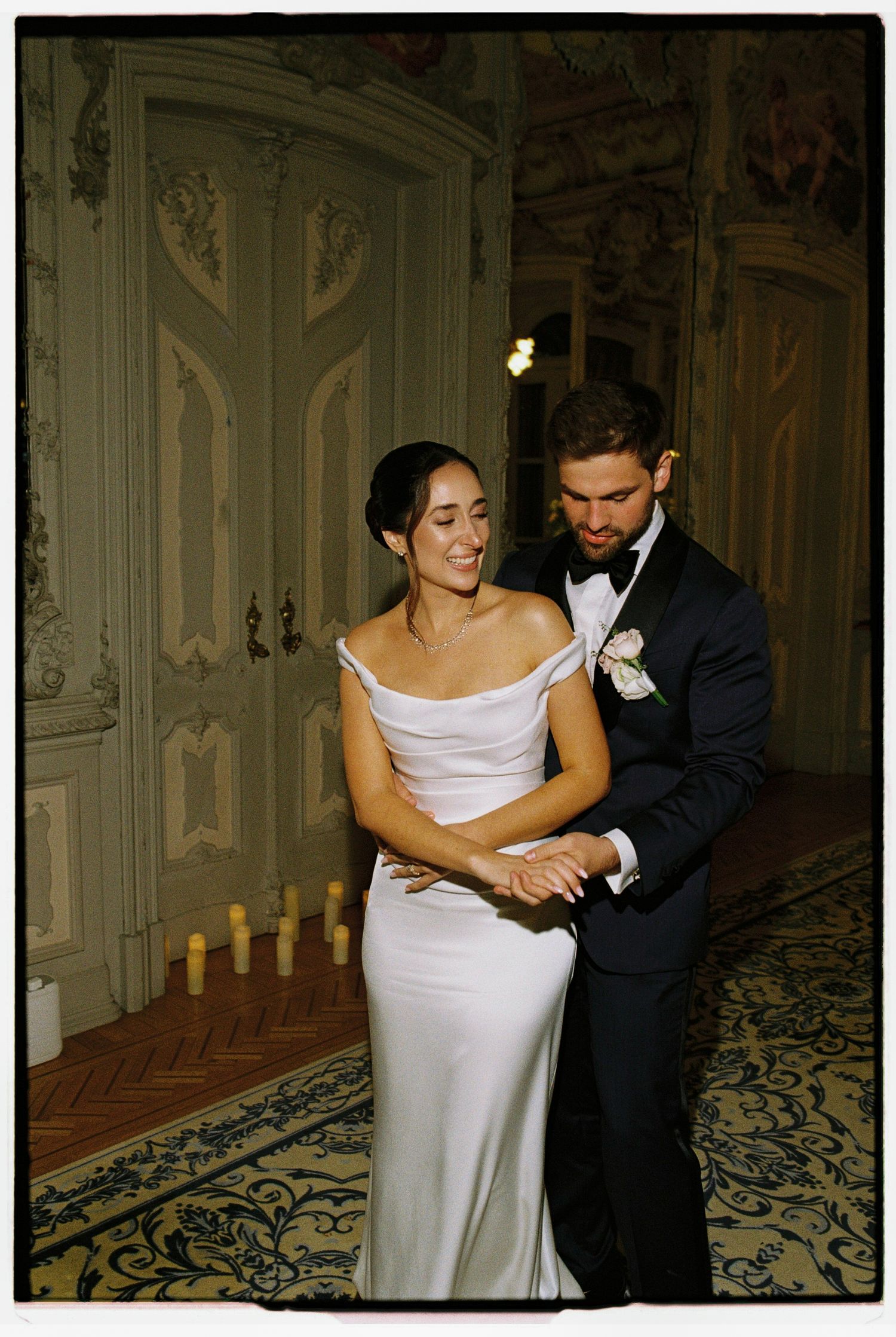 Bride and groom share first dance in elegant ballroom, bride in off-shoulder white gown, groom in navy tuxedo.