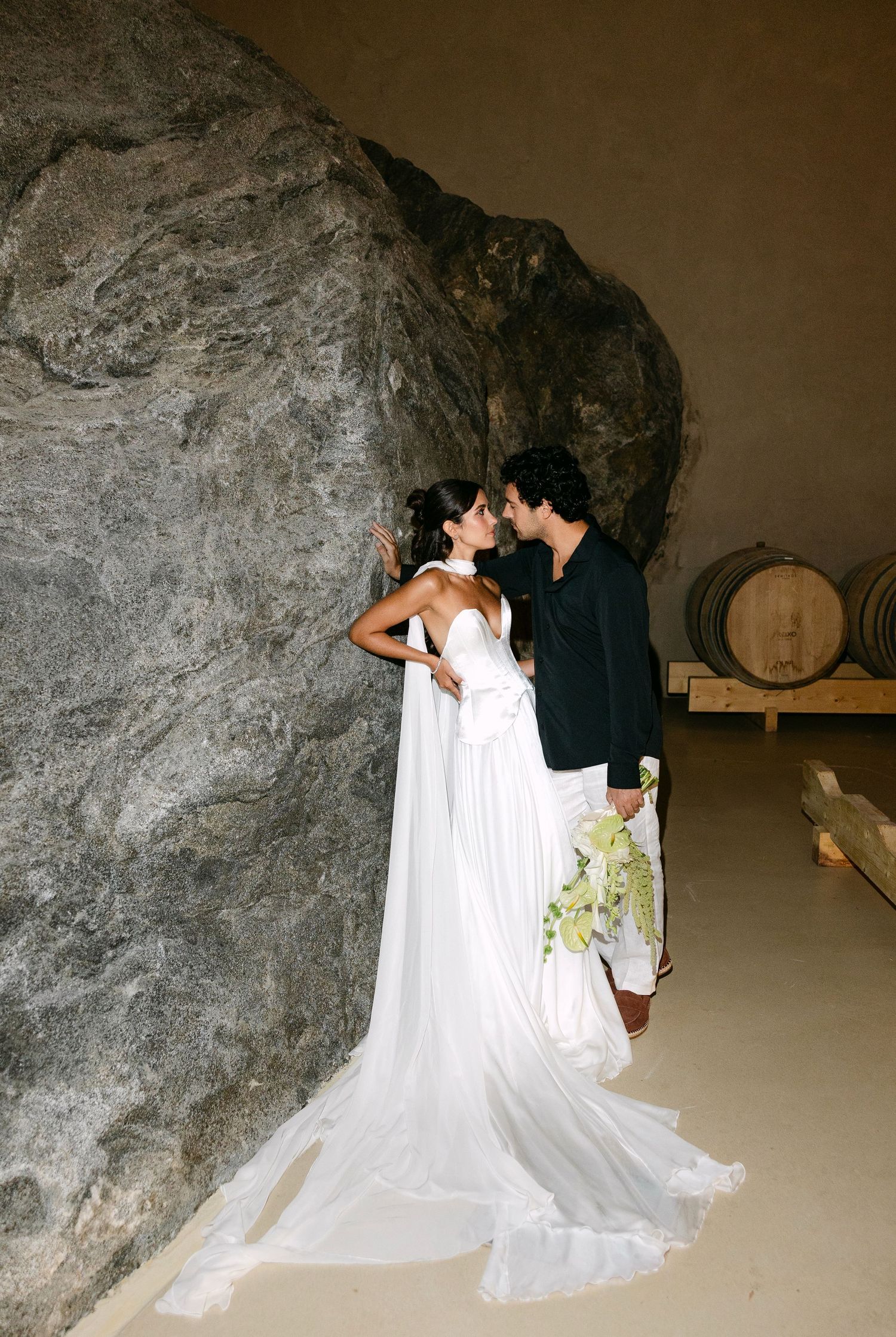 Bride and groom share a kiss beside a large rock in a rustic winery cave with barrels.