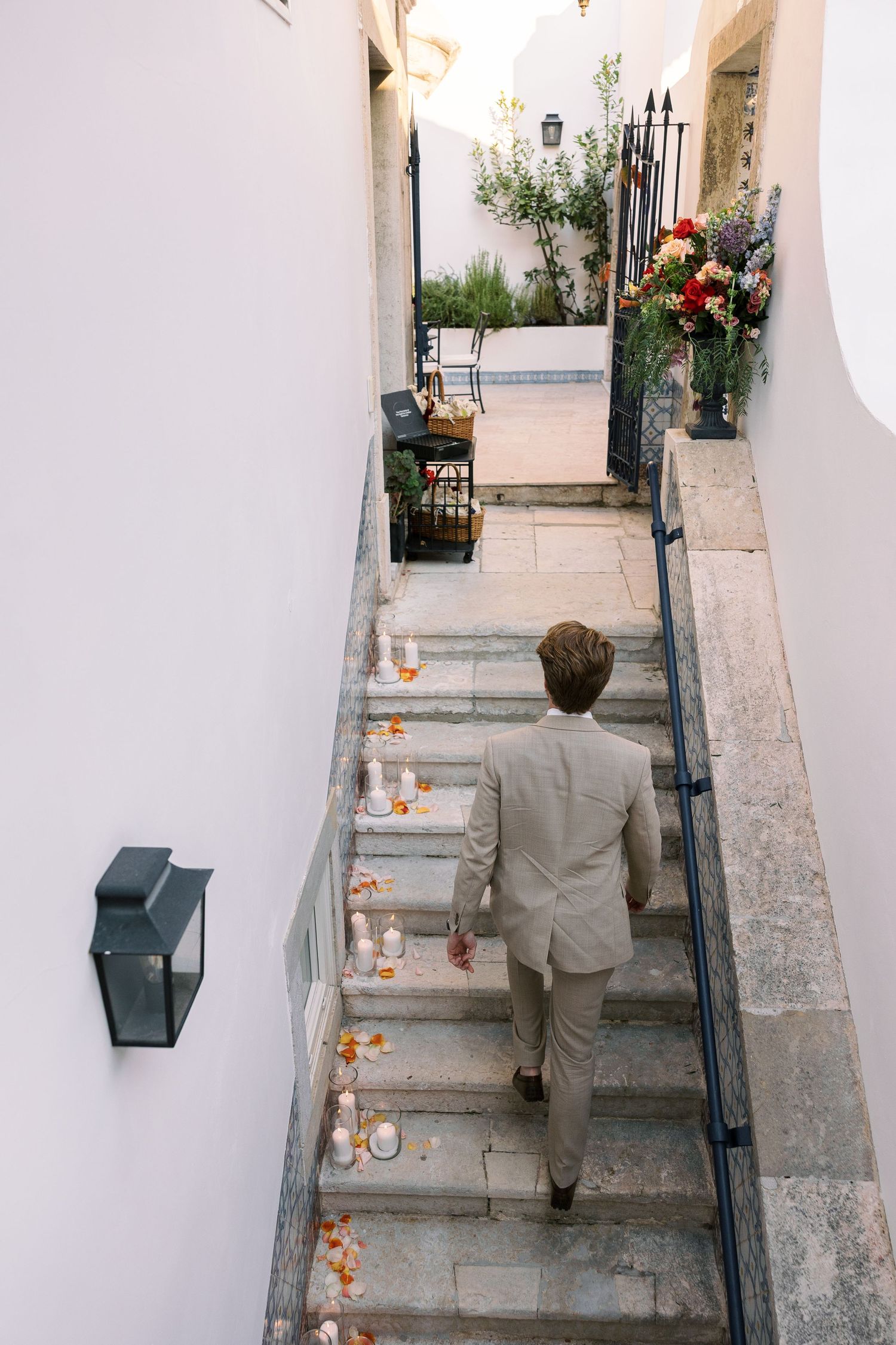 Man in beige suit walking up stone stairs decorated with candles and flower petals at an elegant outdoor venue.