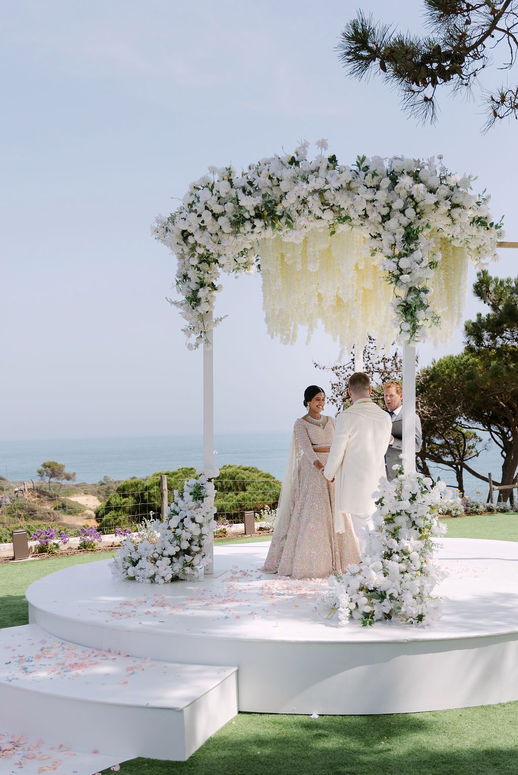 Elegant outdoor wedding ceremony setup with white floral arch and ocean view backdrop.