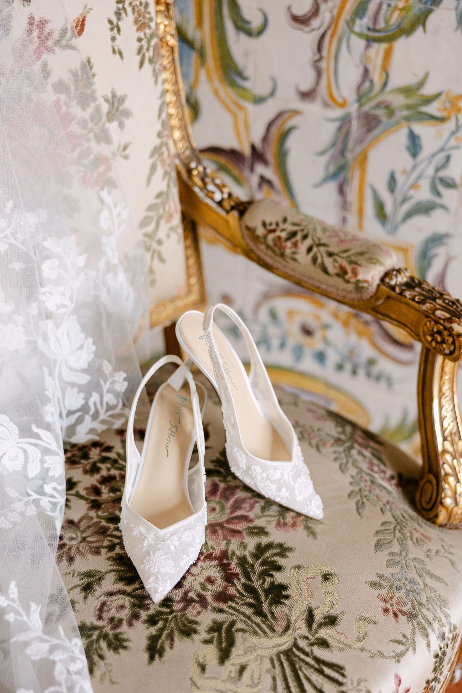 White lace bridal shoes with delicate embroidery details photographed against floral wallpaper.