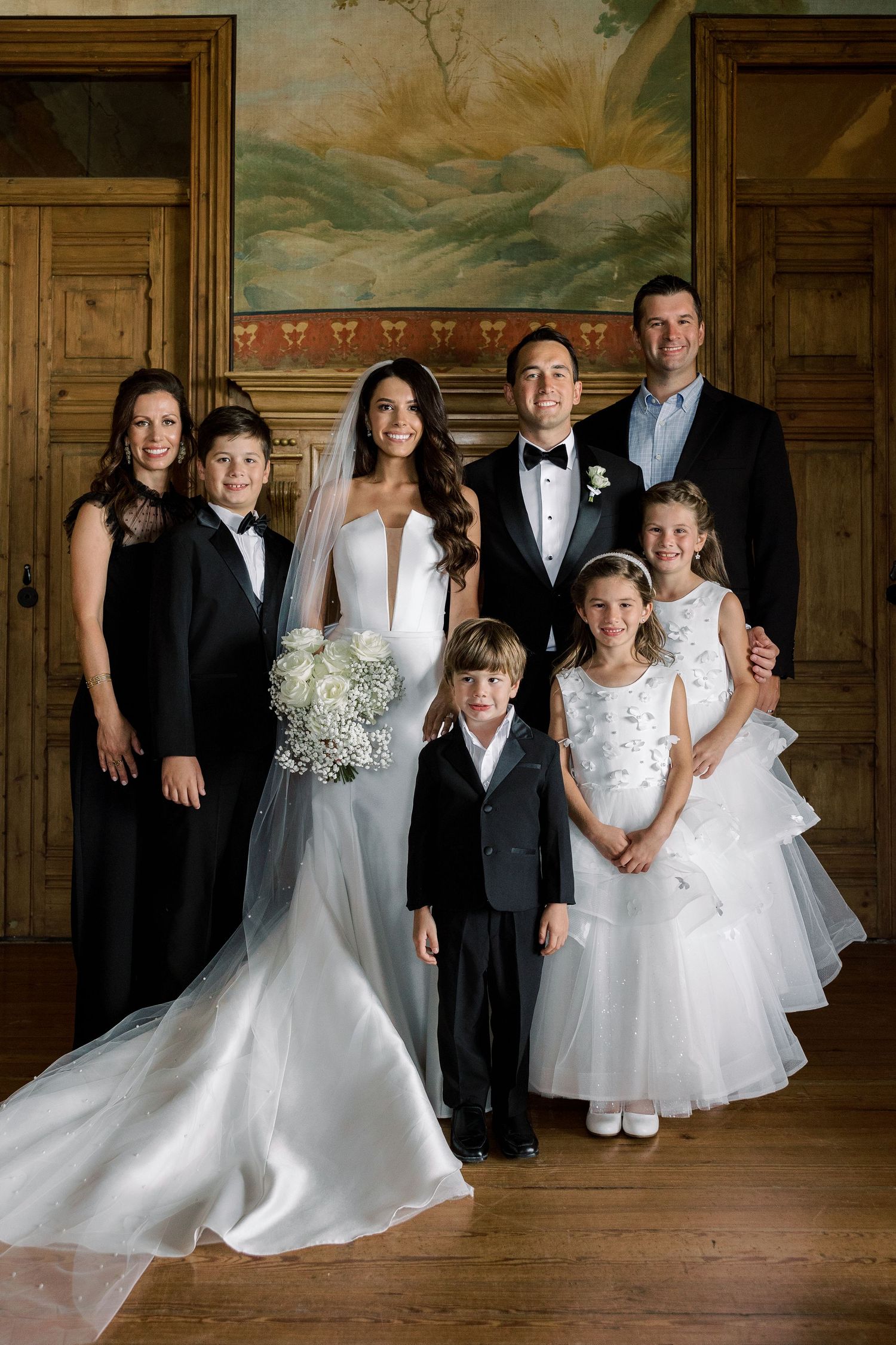 Wedding party in formal attire poses in an elegant room with ornate wall murals and wooden floors.