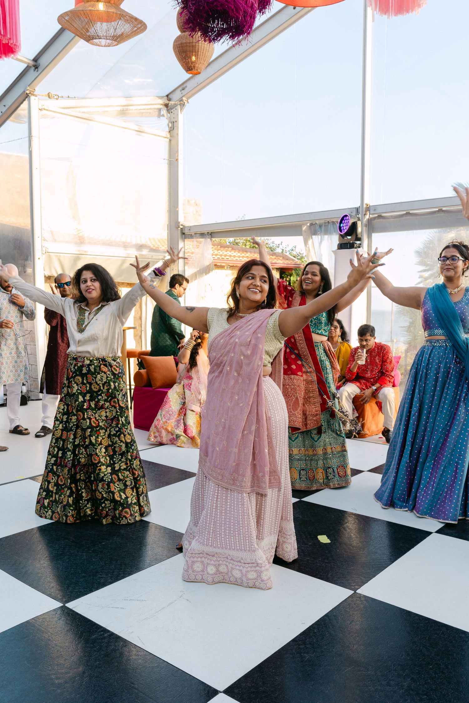 Wedding celebration with guests dancing in colorful Indian dresses on a black and white checkered dance floor.