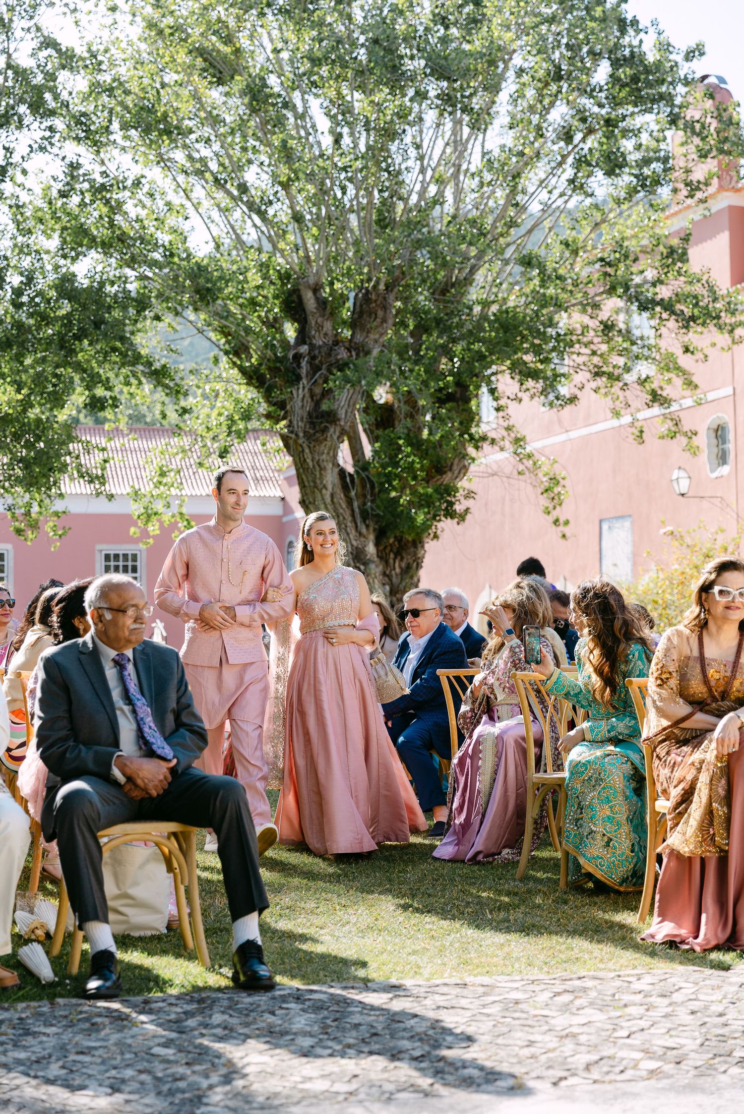Wedding guests in pink and pastel attire gather under trees at an outdoor celebration.