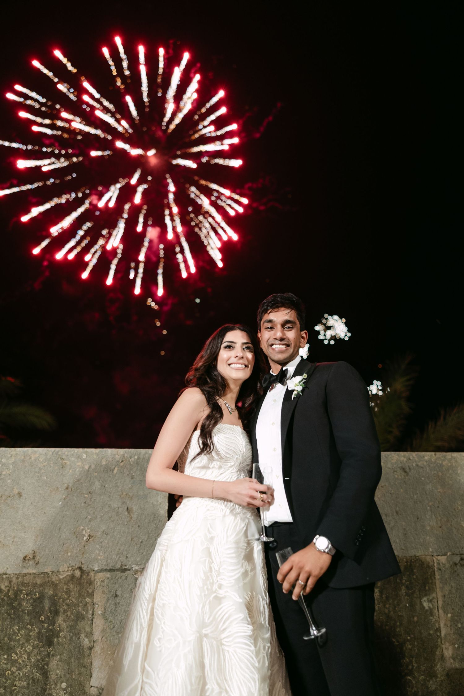 Newlyweds share a romantic moment under red fireworks against a night sky.