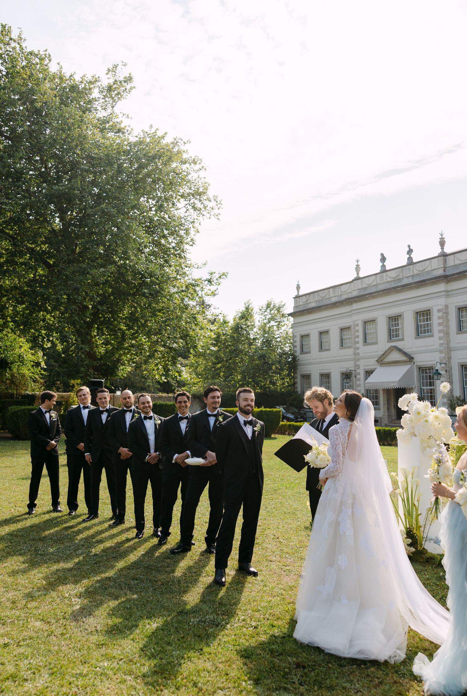 Wedding party poses for formal photos on a grand estate lawn.