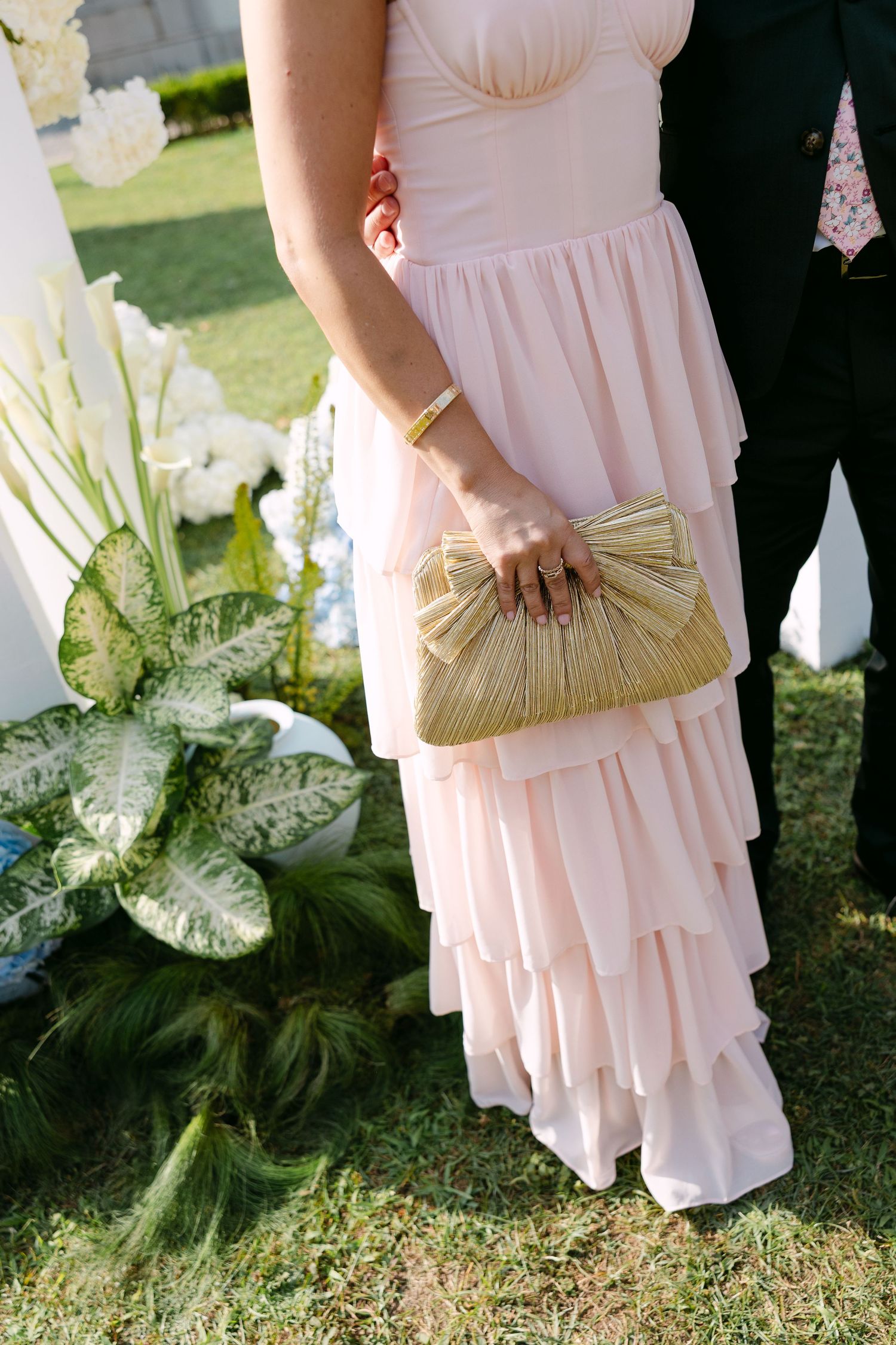 Bridesmaid in blush pink dress holding gold clutch purse against floral arrangement.