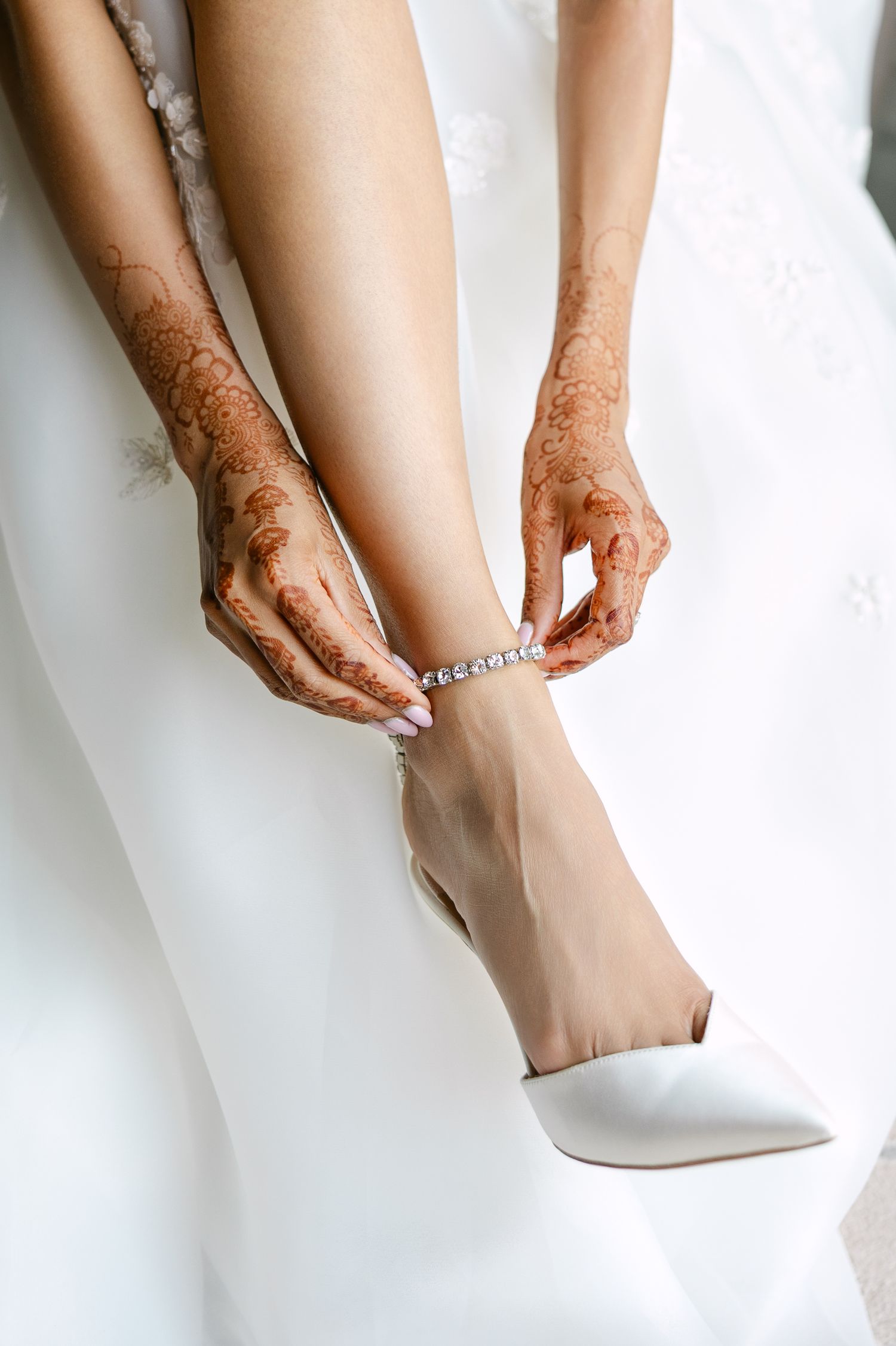 Close-up of henna-decorated hands fastening an anklet with white wedding shoes.